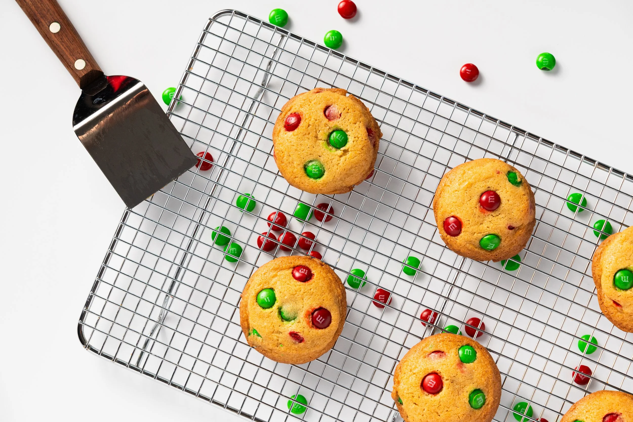 Cookies with red and green M&M candies on a cooling rack, surrounded by scattered M&M candies, with a metal spatula on a white surface.