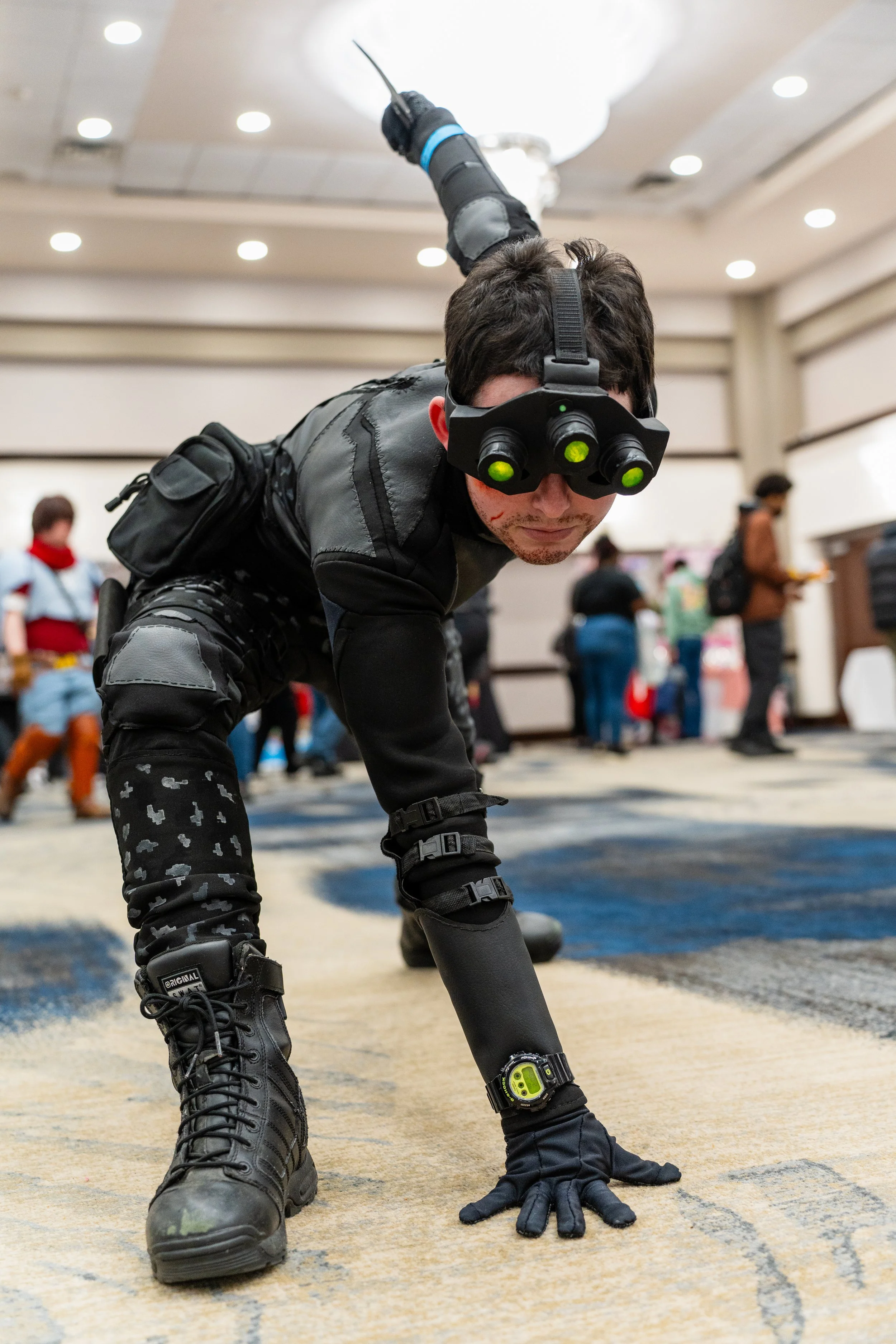 Person dressed in tactical gear with a headlamp and magnifying goggles, crawling on hands and knees on a patterned carpet in a crowded indoor convention or event space.