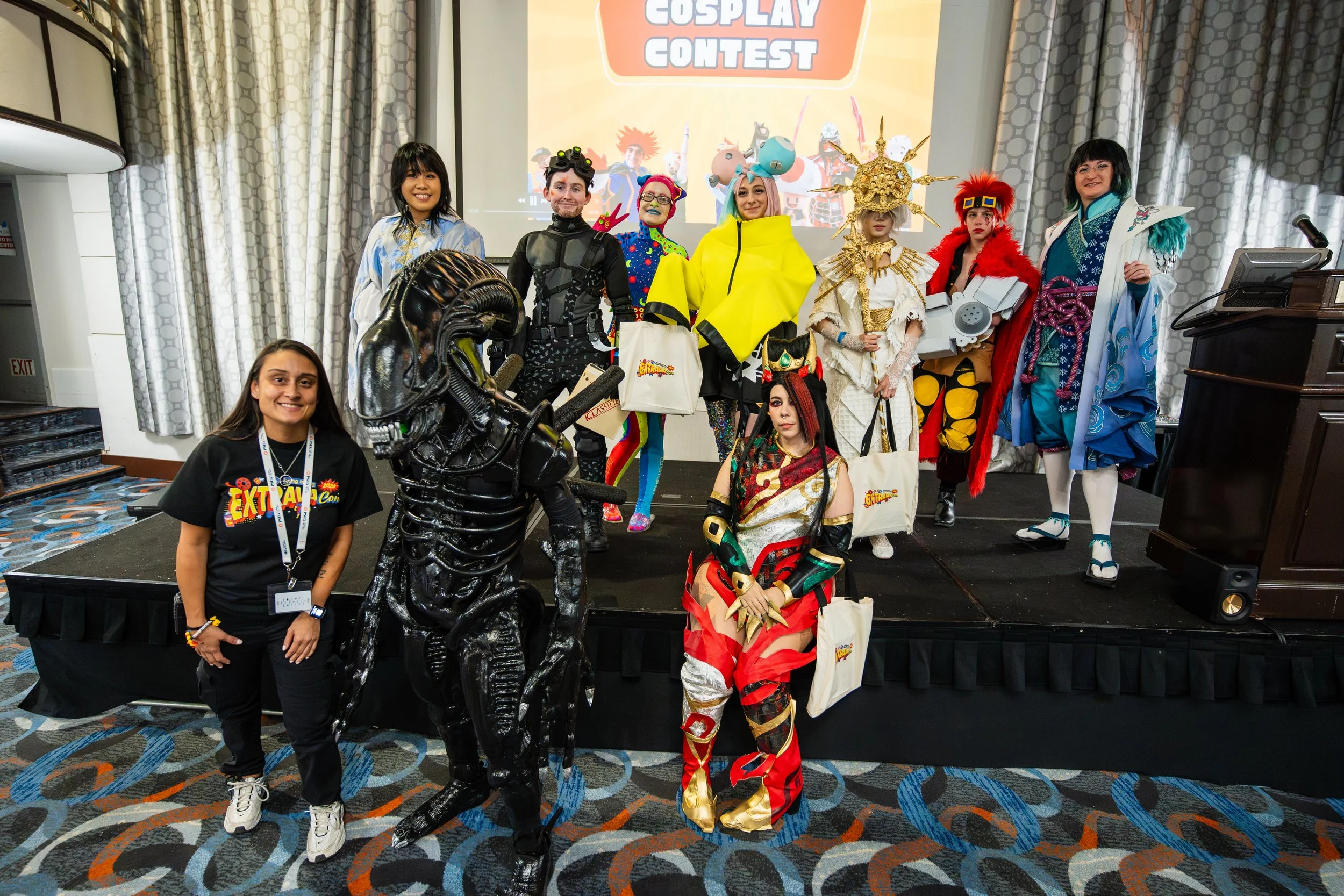 Group of ten people in elaborate cosplay costumes on stage at a cosplay contest, with one person in the front kneeling and others standing behind, backdrop with 'Cosplay Contest' sign, and patterned carpet in the venue.