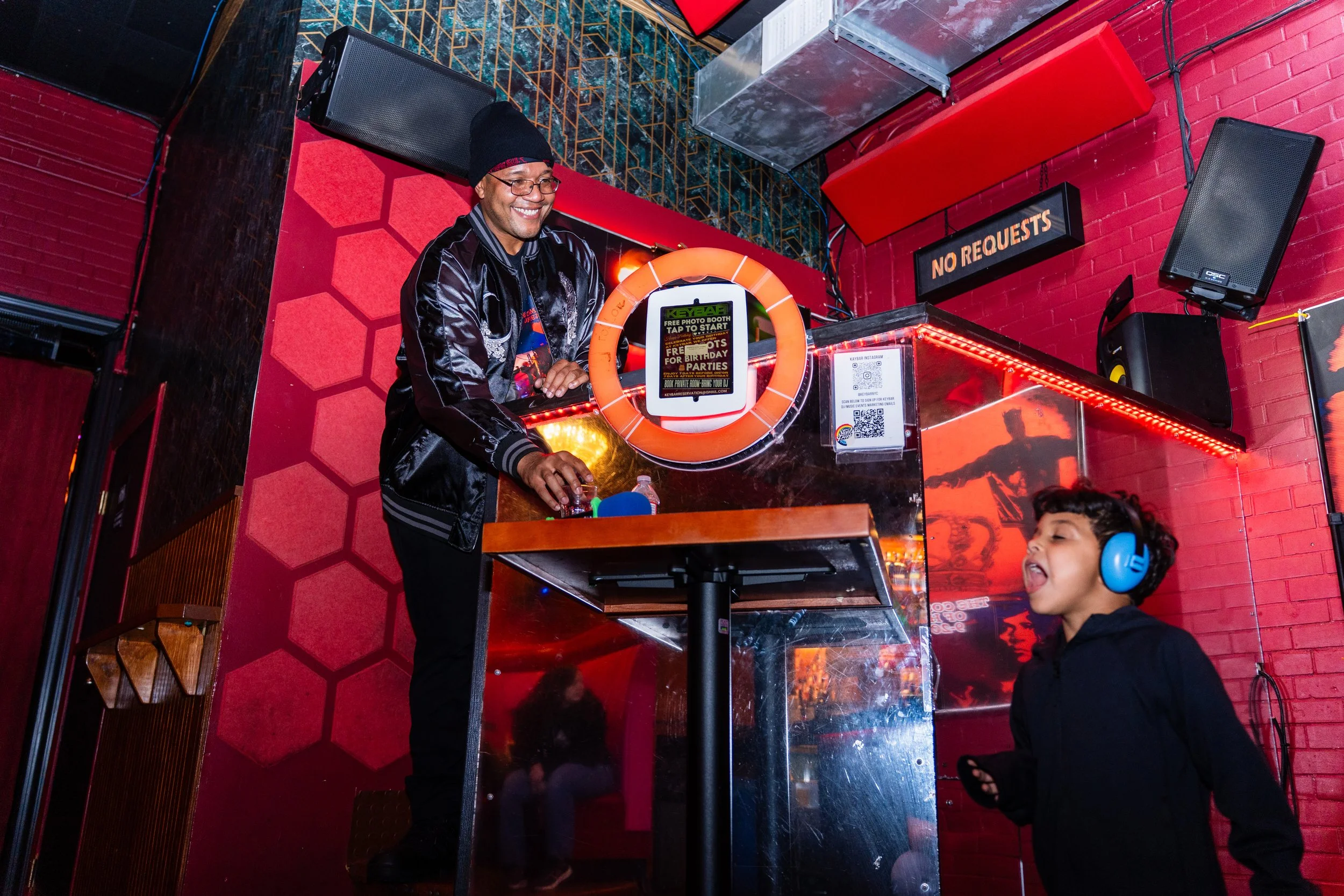 A man wearing a black jacket, beanie, and glasses is smiling behind a DJ booth in a vibrant, red-themed bar or club. A young girl with headphones and black hoodie is singing or talking into a microphone, looking excited.