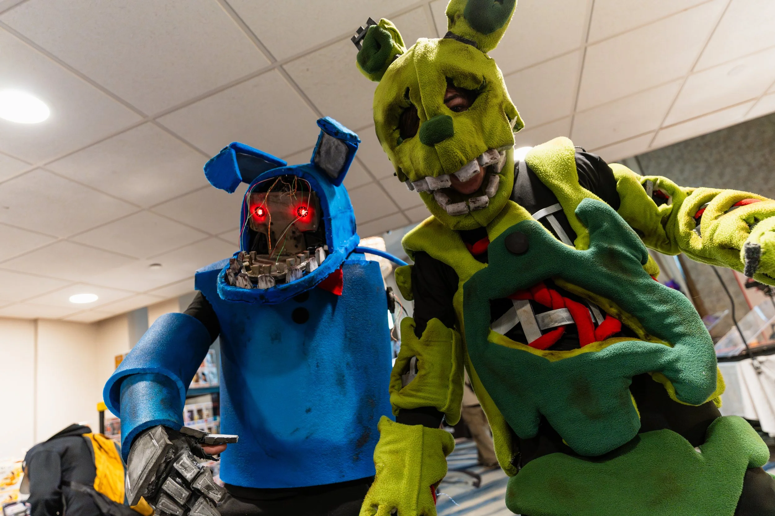 Two people in elaborate costumes, one as an animatronic-style robot with glowing red eyes and blue body, and the other as a large, green, monster-like character with exaggerated facial features, both posing inside a room with a ceiling and shelves in
