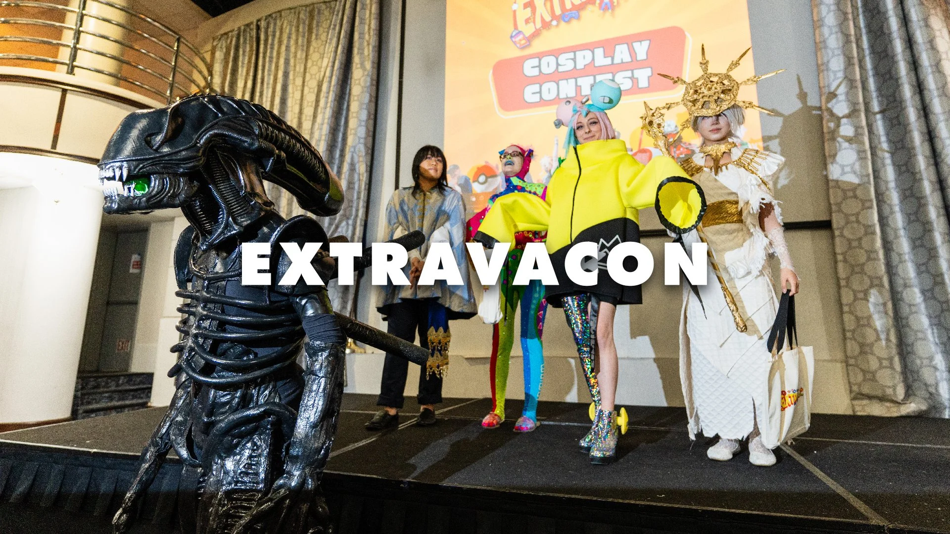 People in colorful costumes on a stage at an event, with a large screen behind them displaying the words 'ExtravaCon' and 'Cosplay Contest'.