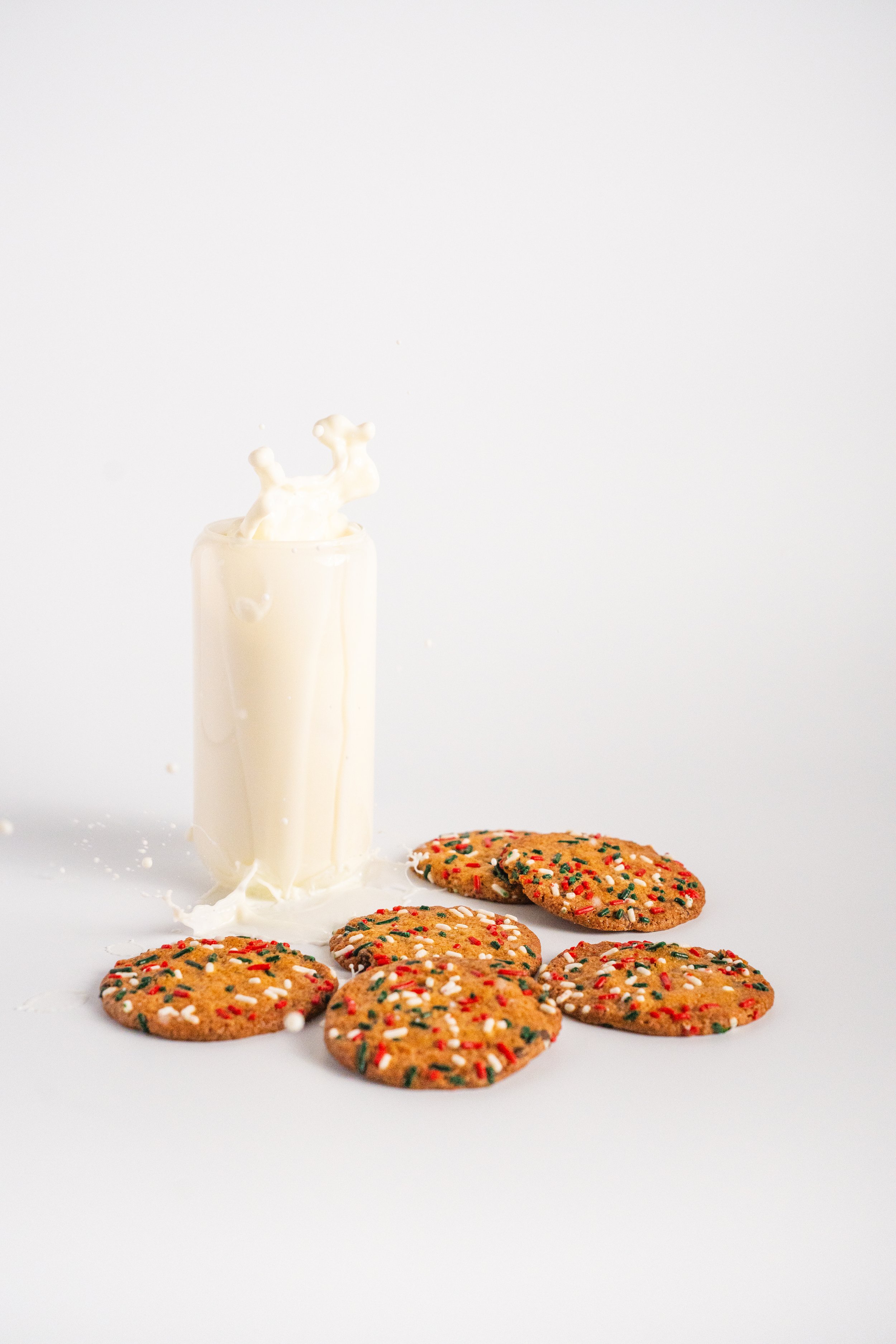 A glass of milk with a splash on the side, surrounded by five sugar cookies decorated with red, green, and white sprinkles on a white background.