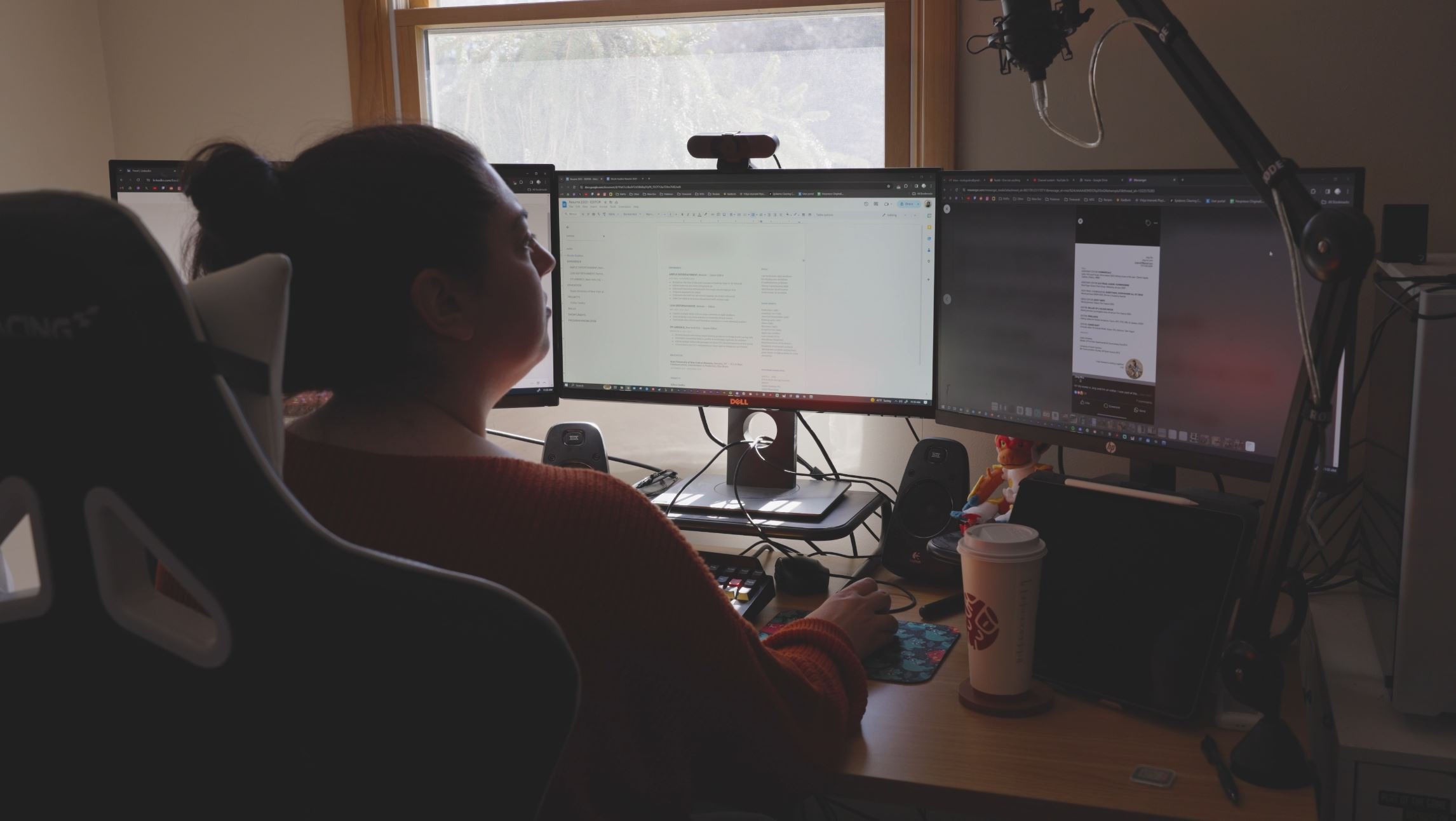 A woman working at a desk with three computer monitors, a drink cup, and a robotic arm near her.