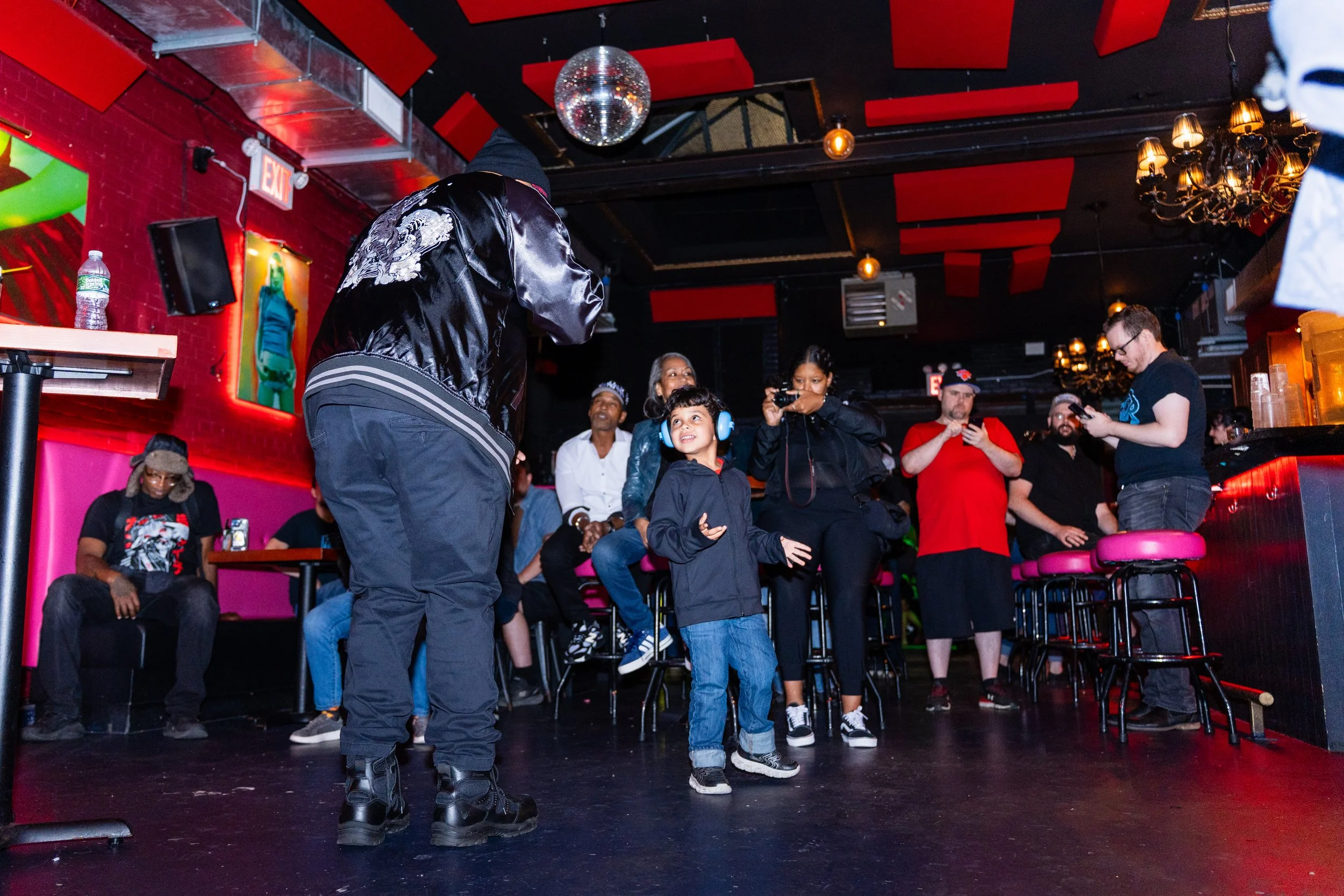 A young boy wearing headphones dancing on the stage of a nightclub with a group of men and women watching and taking photos.