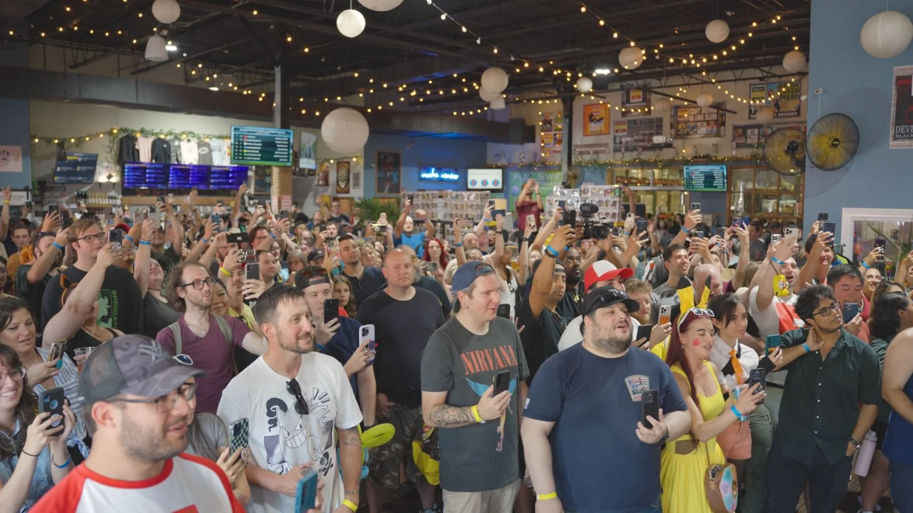 Large crowd of people at an indoor event, many holding up smartphones and cameras to take photos or videos, with decorative string lights and lanterns overhead.