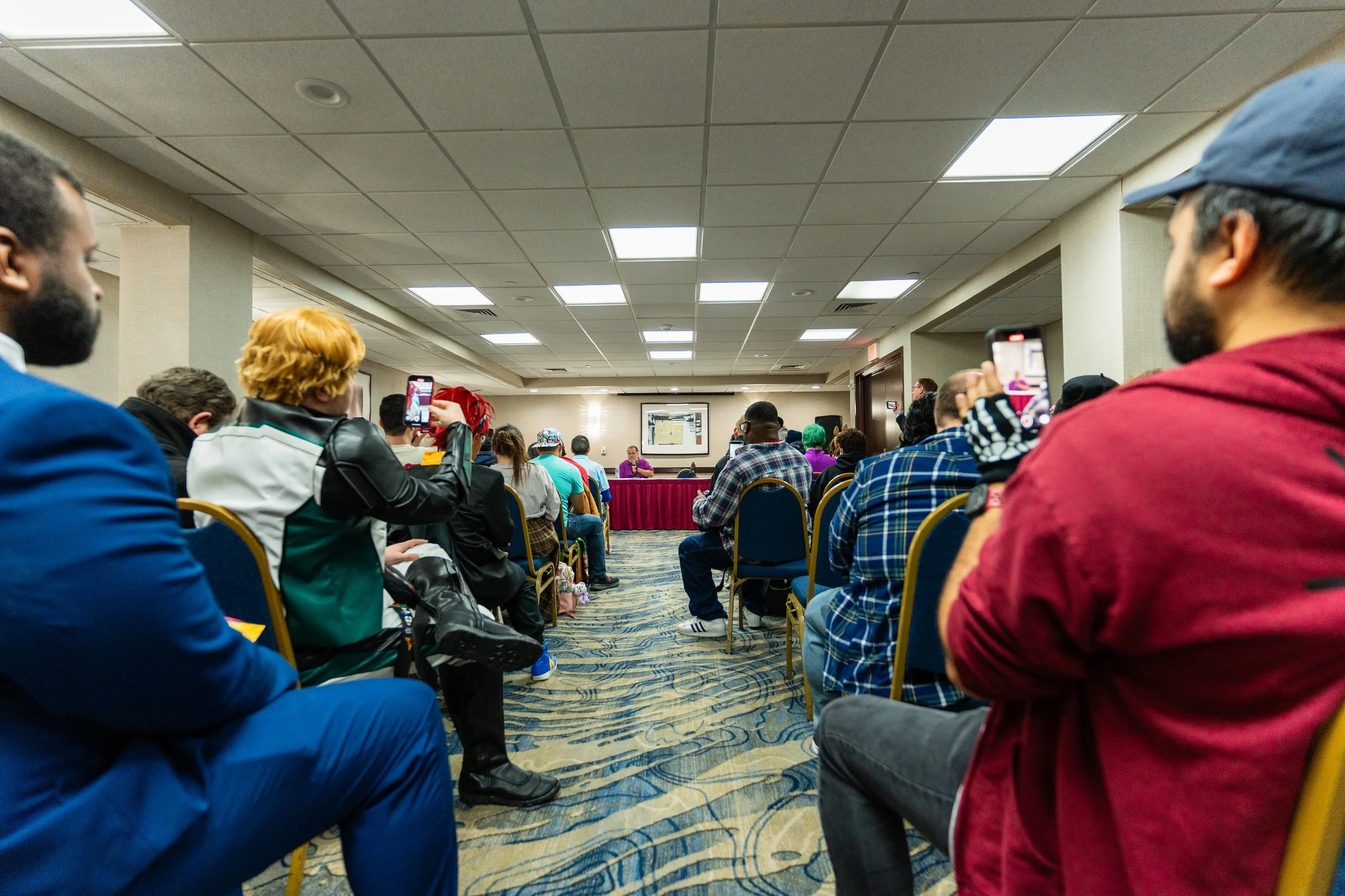 People attending a conference in a large room, some are taking photos or videos of the speaker at the front of the room.