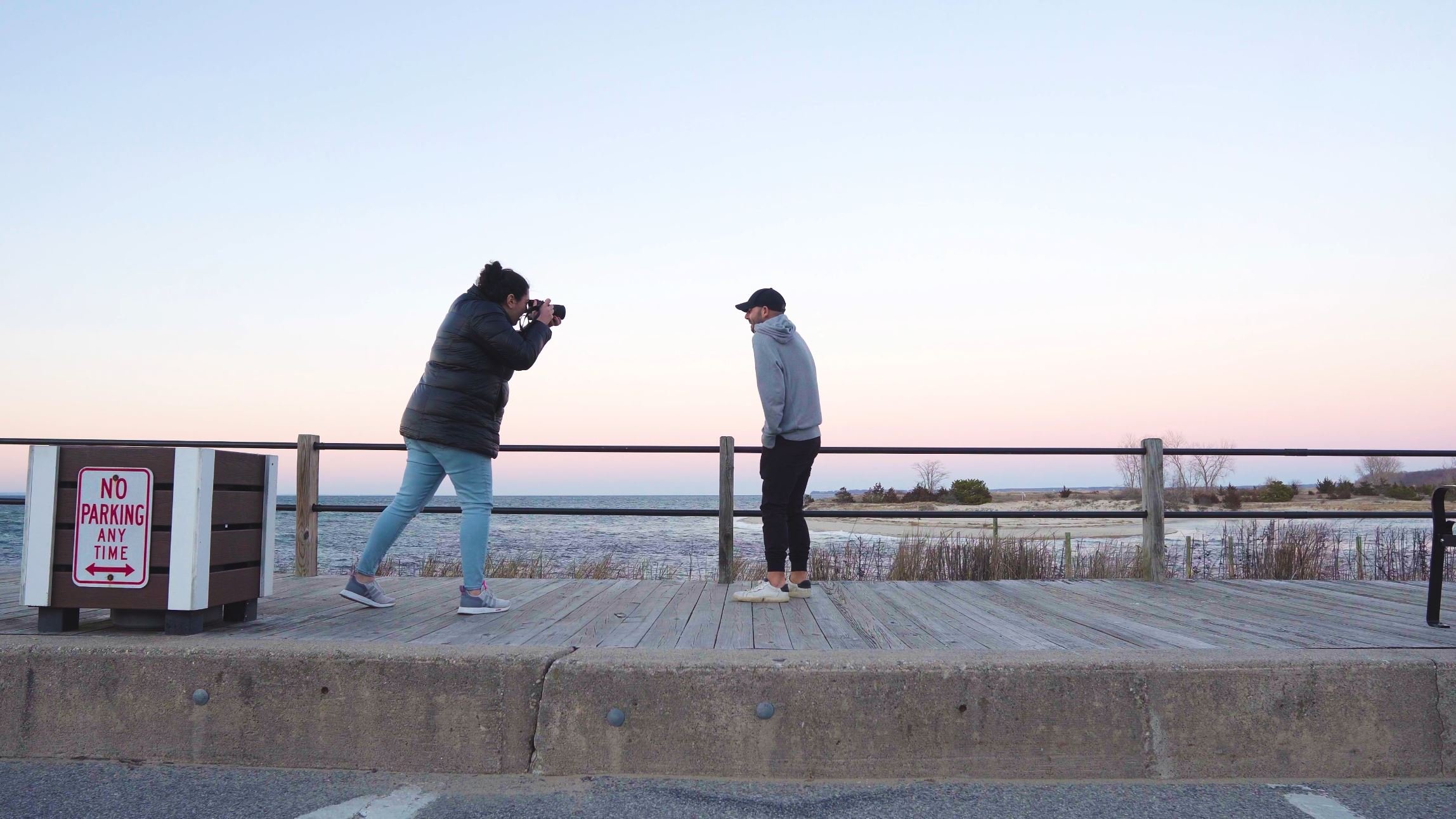 A woman taking a photograph of a man standing on a wooden boardwalk near a body of water at sunset or sunrise.