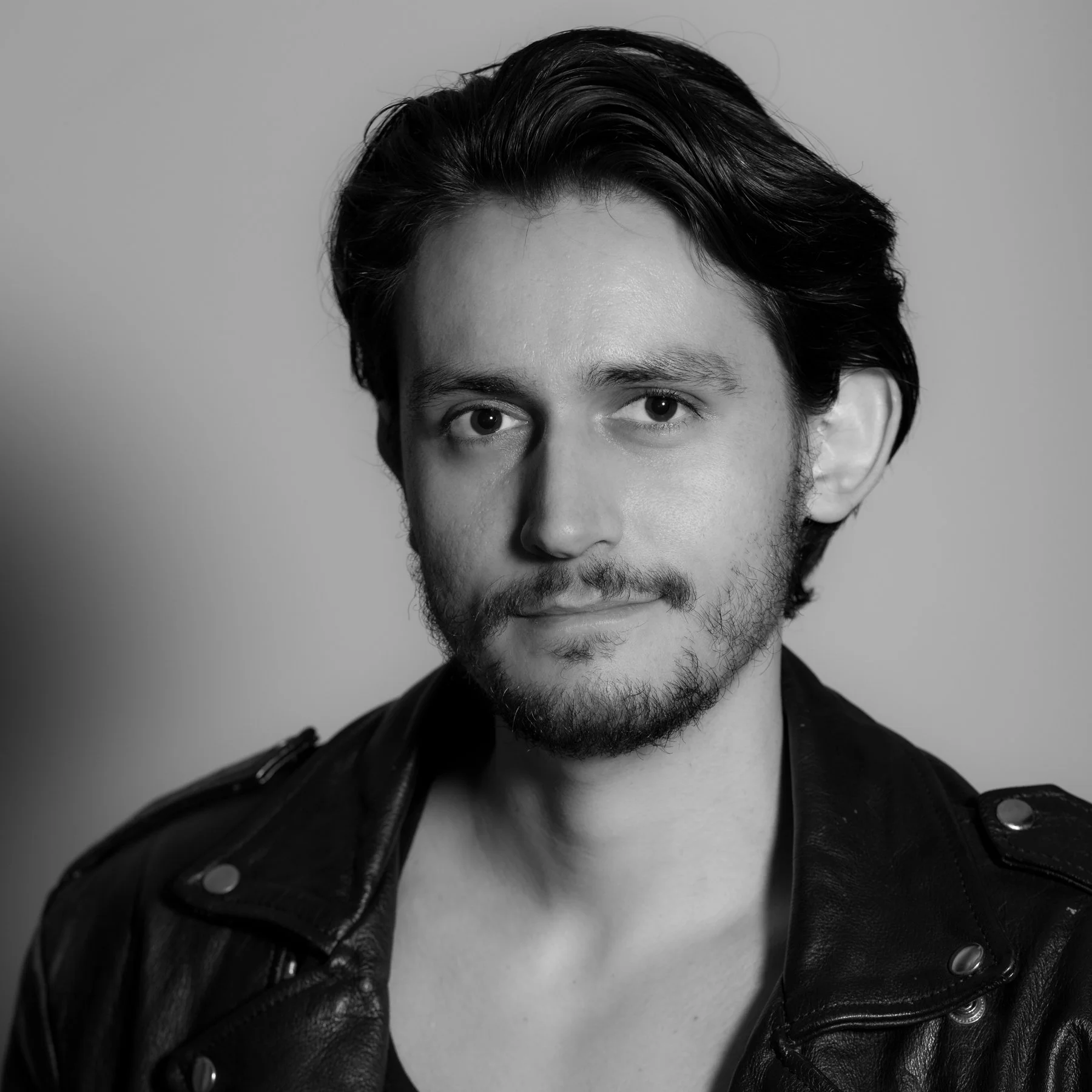 Close-up portrait of a man with dark hair and a beard, wearing a black leather jacket with metal buttons, looking directly at the camera against a plain light gray background.