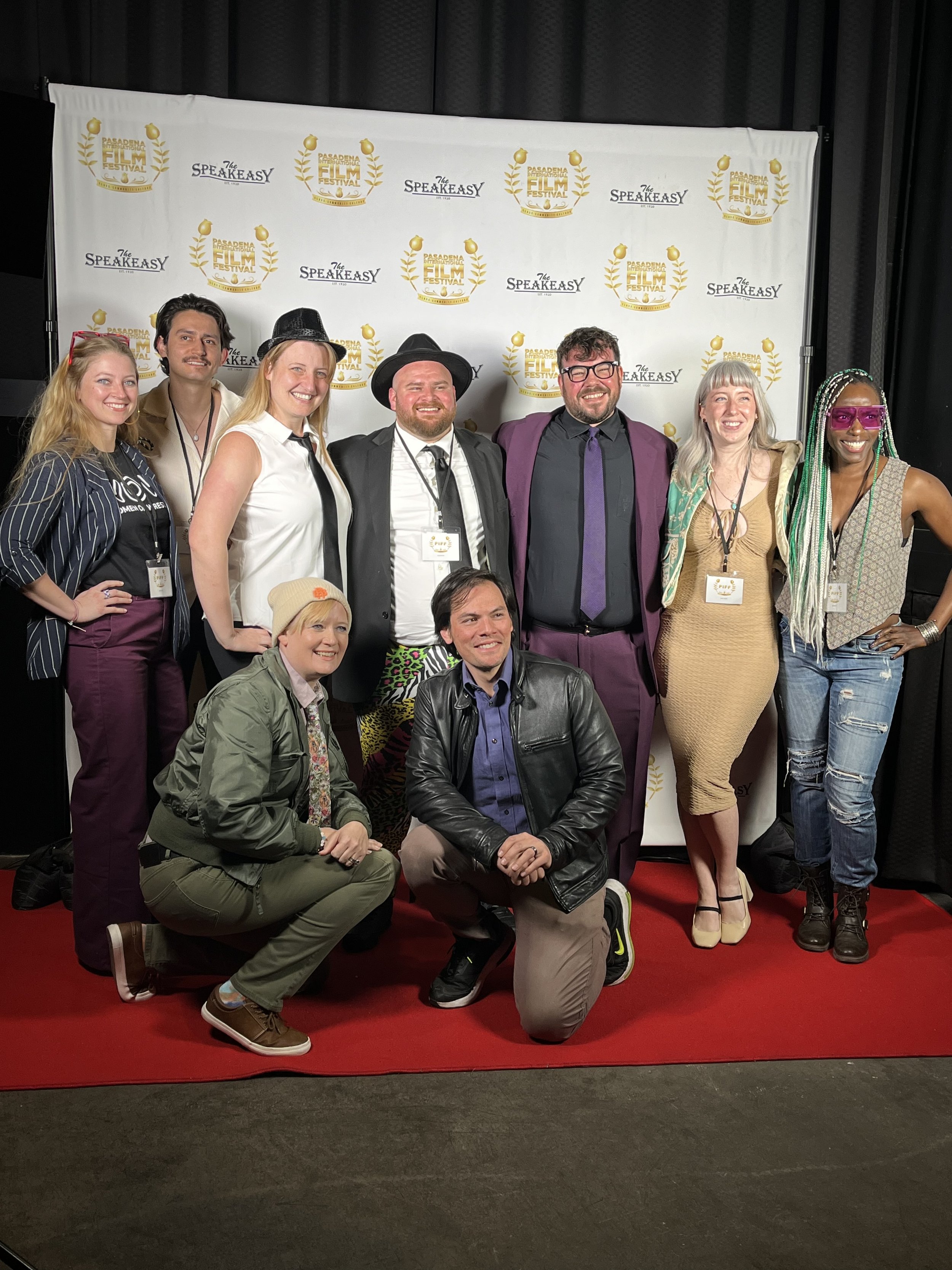 Group of ten diverse people posing on red carpet at Pasadena International Film Festival, standing in front of a white backdrop with festival logos, some standing and some kneeling, smiling.