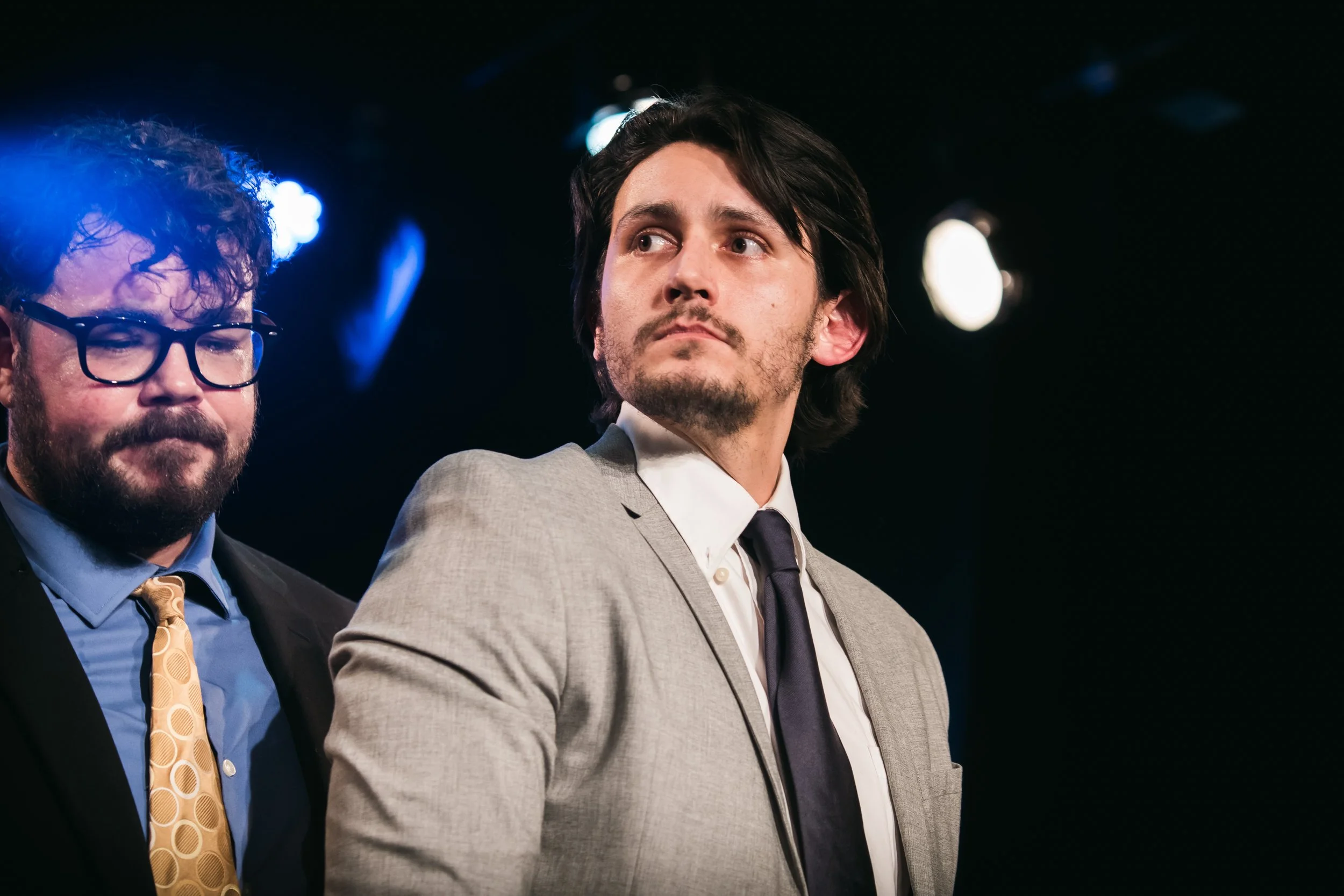 Two men dressed in suits, one with dark hair and the other with glasses and curly hair, standing on a stage with dark background and bright stage lights.
