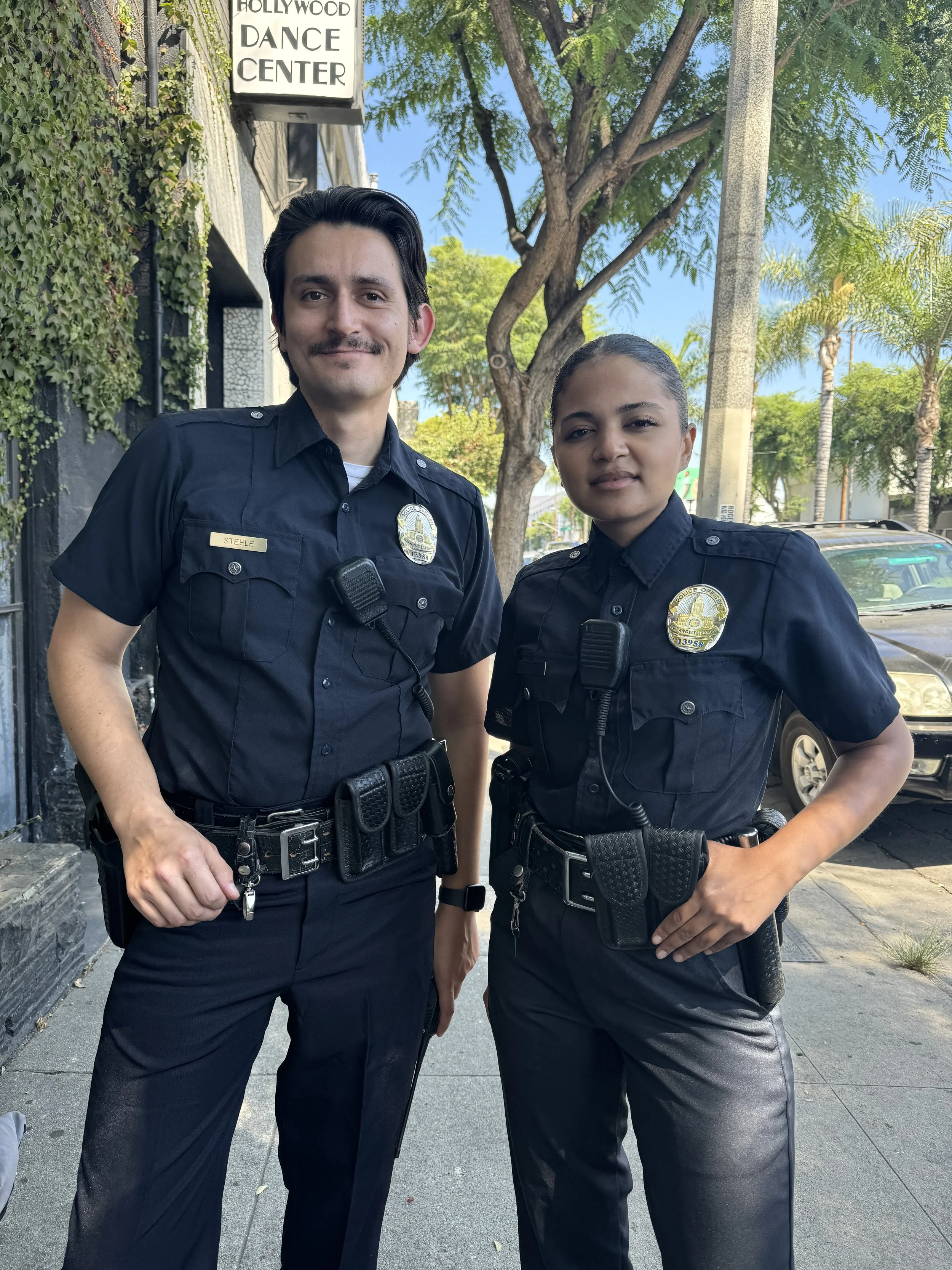 Two police officers, a man and a woman, standing outside in front of a tree and a building with a sign that reads 'Hollywood Dance Center'. They are wearing dark uniforms with badges and police equipment.