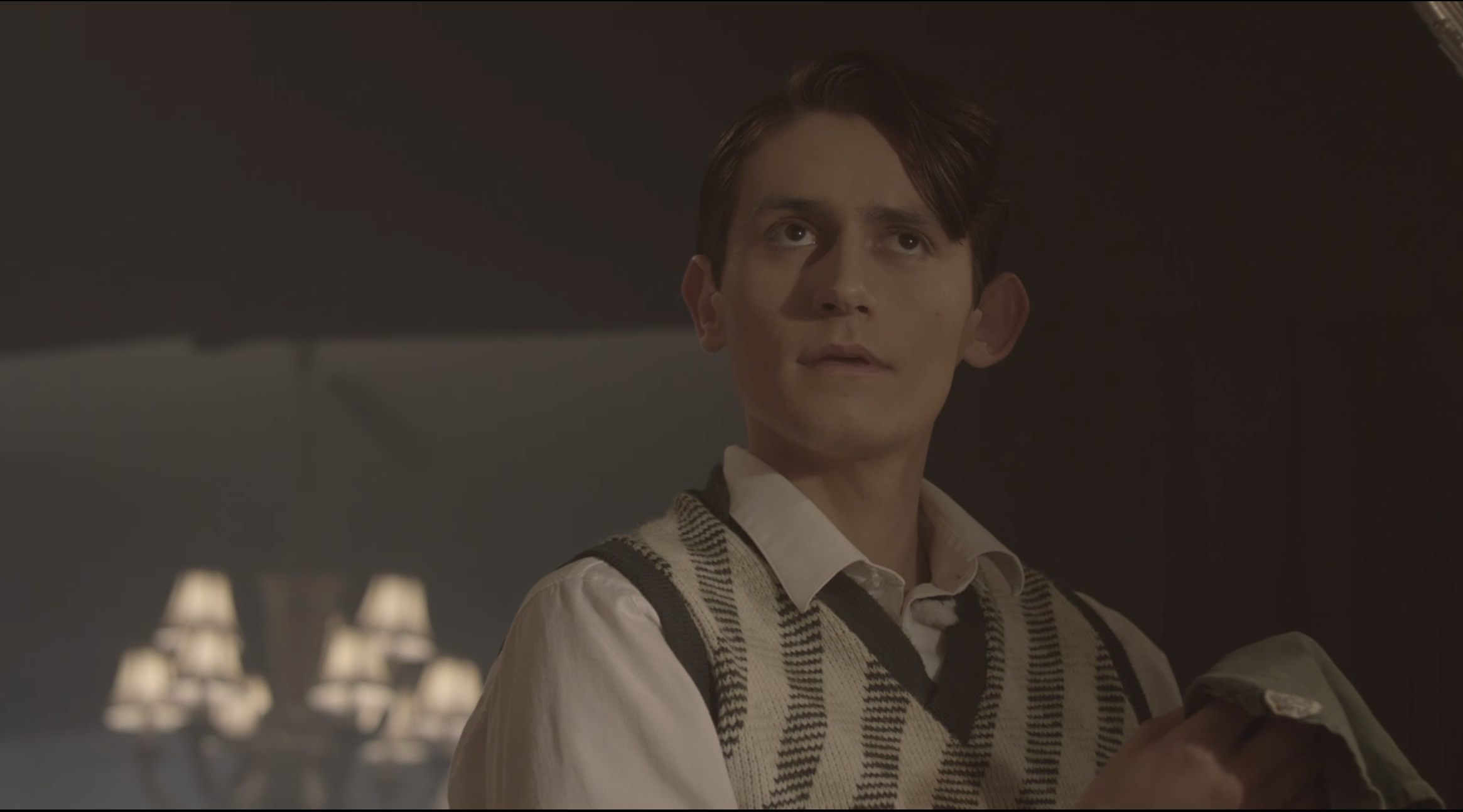 a Young Gavin Velez with brown hair looking upwards, wearing a checkered vest over a white shirt in a dimly lit room with hanging lamps.