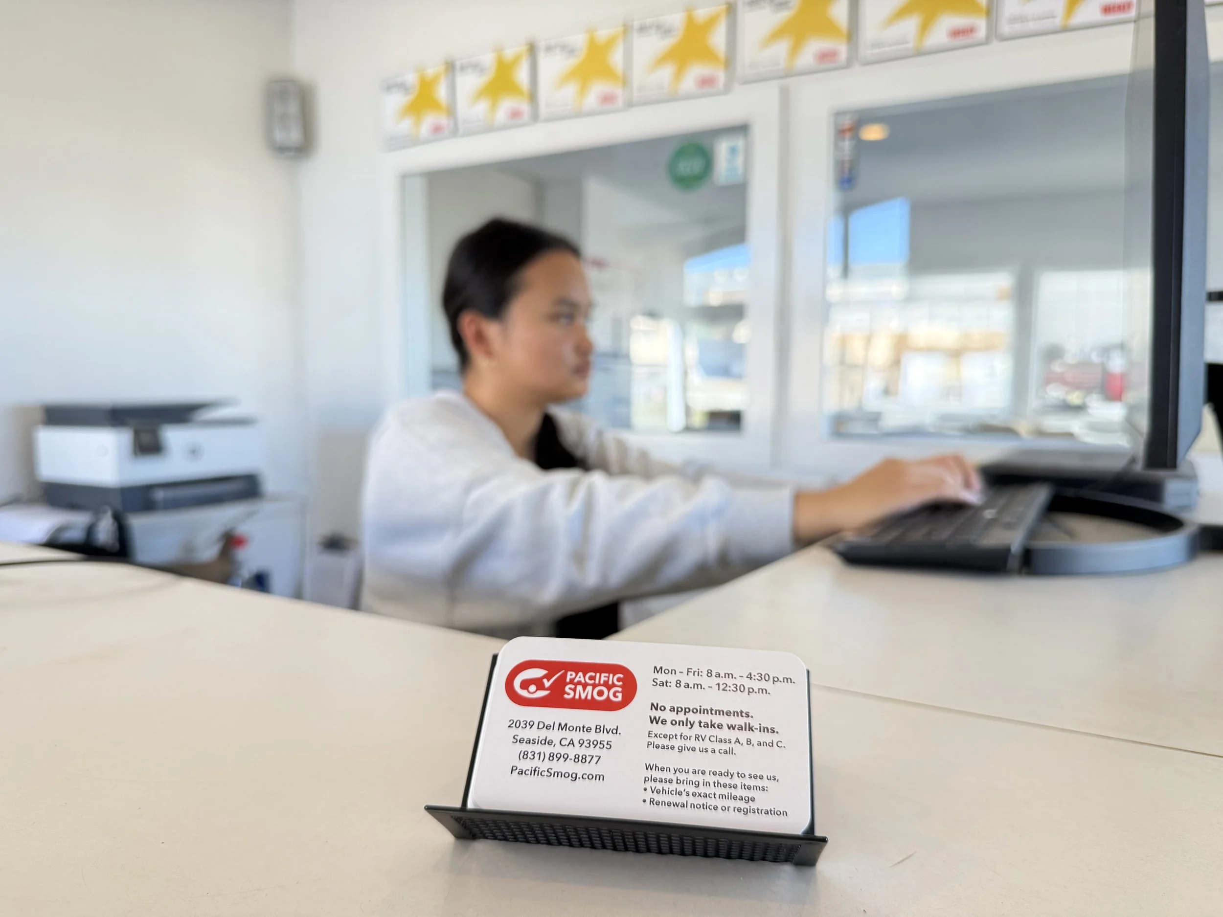 Pacific Smog business card in the foreground with a front desk team member assisting customers in the background. "Best of Monterey County" award plaques are displayed on the wall behind the reception desk.