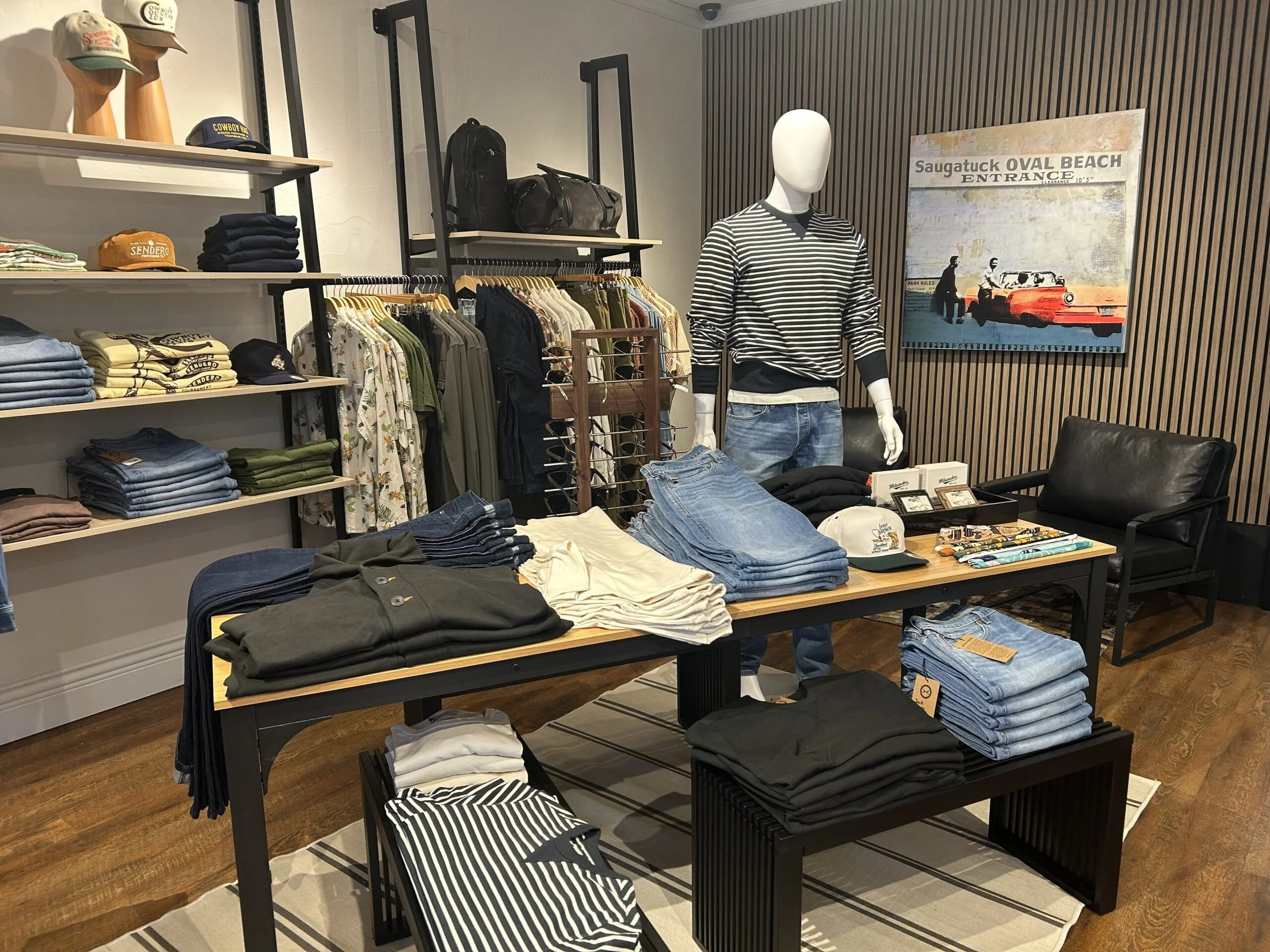 Clothing store display with denim jeans, t-shirts, hats, backpacks, and a mannequin wearing a striped shirt and jeans, with a Saugatuck Oval Beach sign on the wall.