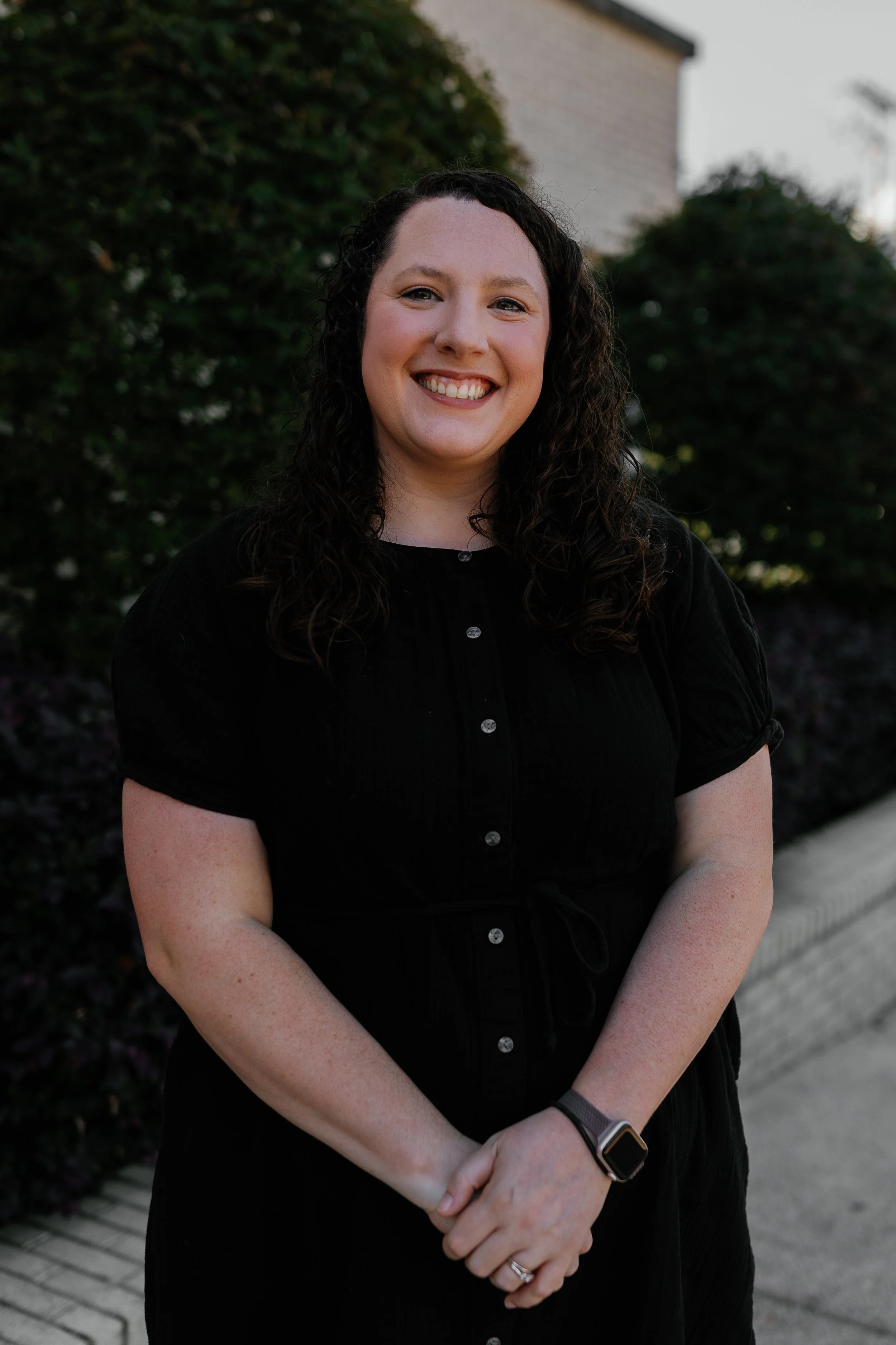A woman with curly brown hair smiling outdoors in a park, wearing a black cardigan over a green top.