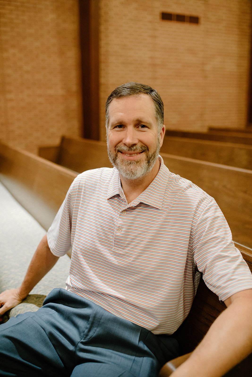 A smiling man with a beard and mustache, wearing a striped polo shirt, sitting on a wooden pew in a church or similar setting.
