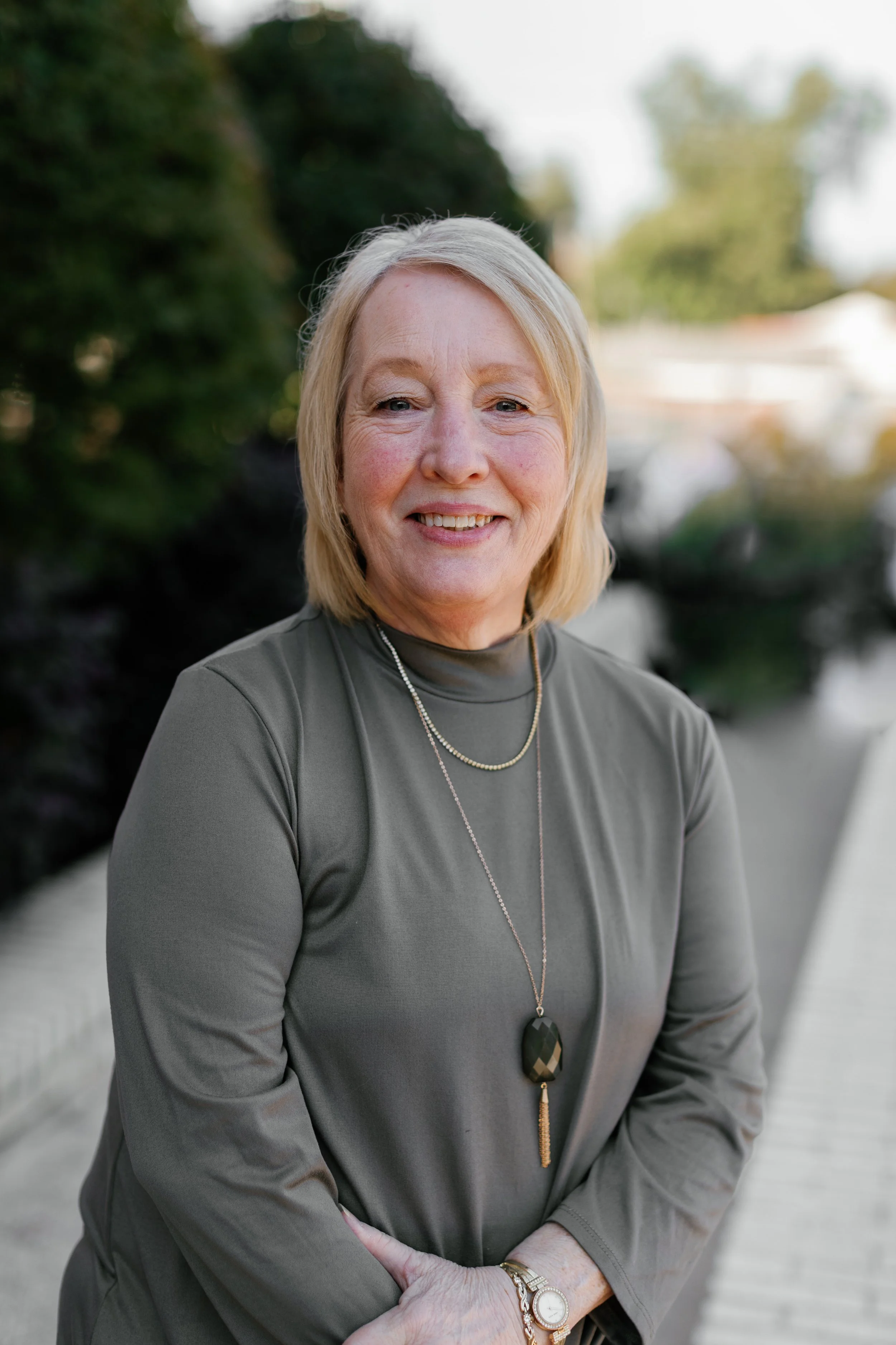 A smiling middle-aged woman with strawberry blonde hair, wearing a green button-up shirt and a gold necklace with a cross pendant, standing outdoors near a building and sidewalk.