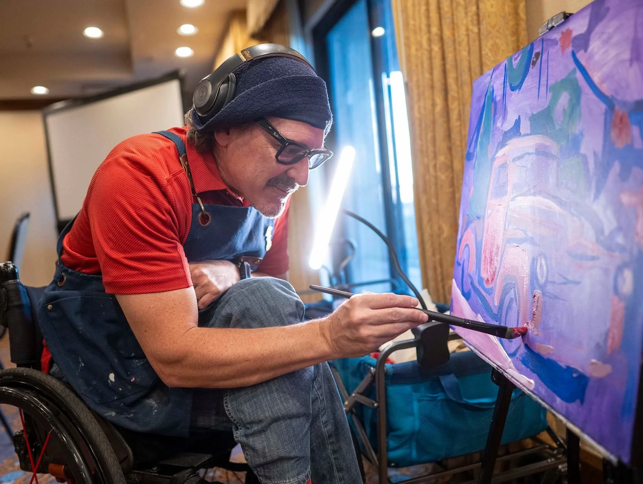 An artist in a wheelchair creating a colorful painting on a large canvas, wearing a blue apron, glasses, a beanie, and a red shirt in an indoor studio setting.