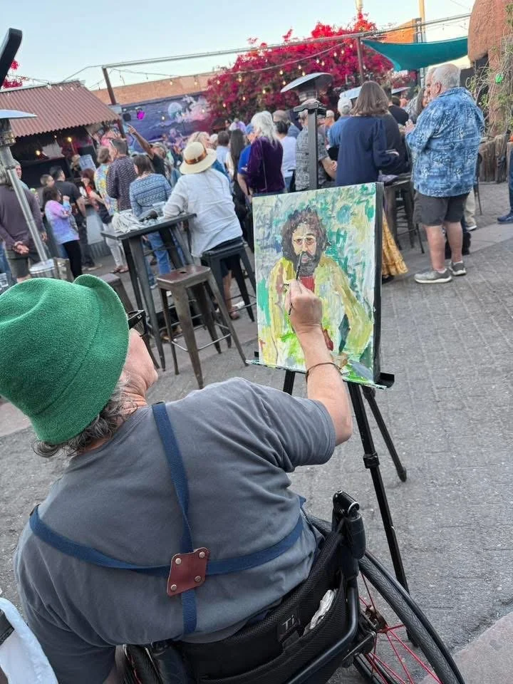 A person in a green beanie and gray shirt is painting a portrait on a canvas set up on an easel at an outdoor event. They are seated in a wheelchair and holding a brush, working on a painting of a man with dark curly hair and glasses. The scene is lively with a crowd of people gathered, some standing around tall tables, in front of a building with a pink flowering tree, and the sky is clear.