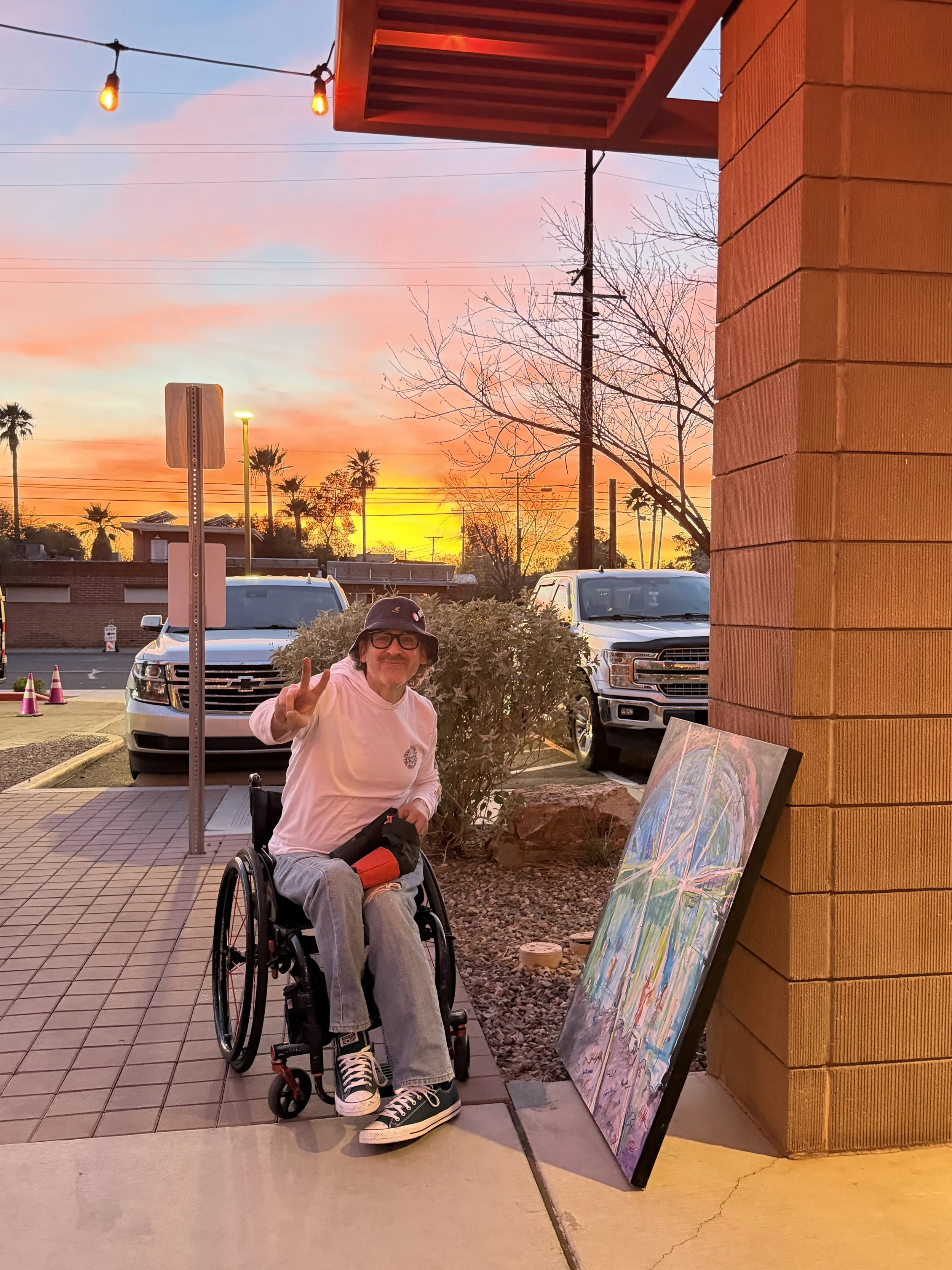 A man in a wheelchair making a peace sign with his fingers, sitting outdoors near a wall with paintings, during sunset with colorful clouds and palm trees in the background.
