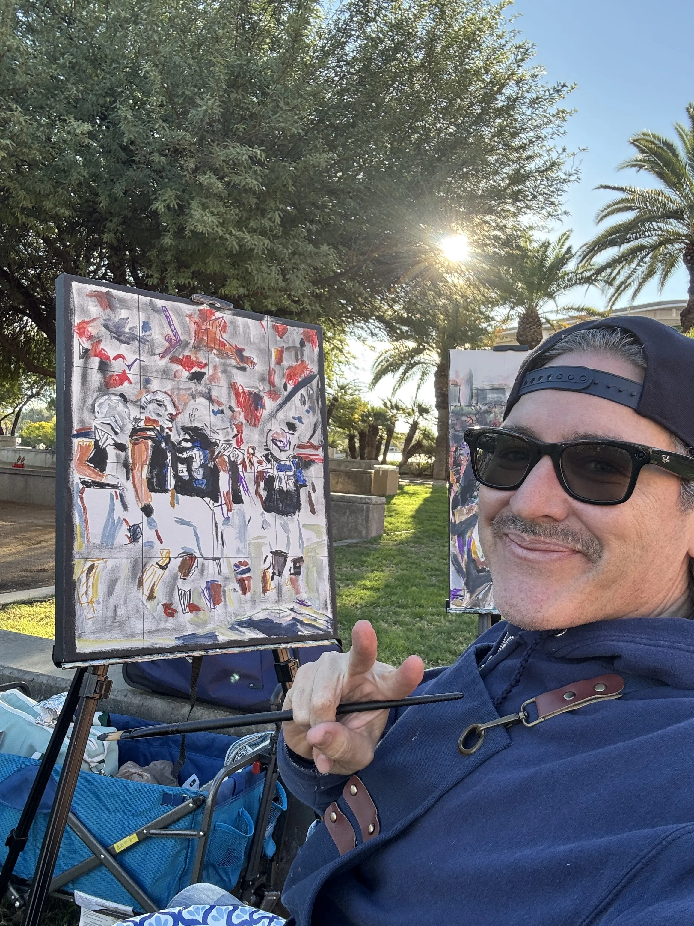 A man with sunglasses and a backwards cap giving a thumbs-up with a smile, standing outdoors near art displayed on easels, with trees and palm trees in the background under a bright sky.