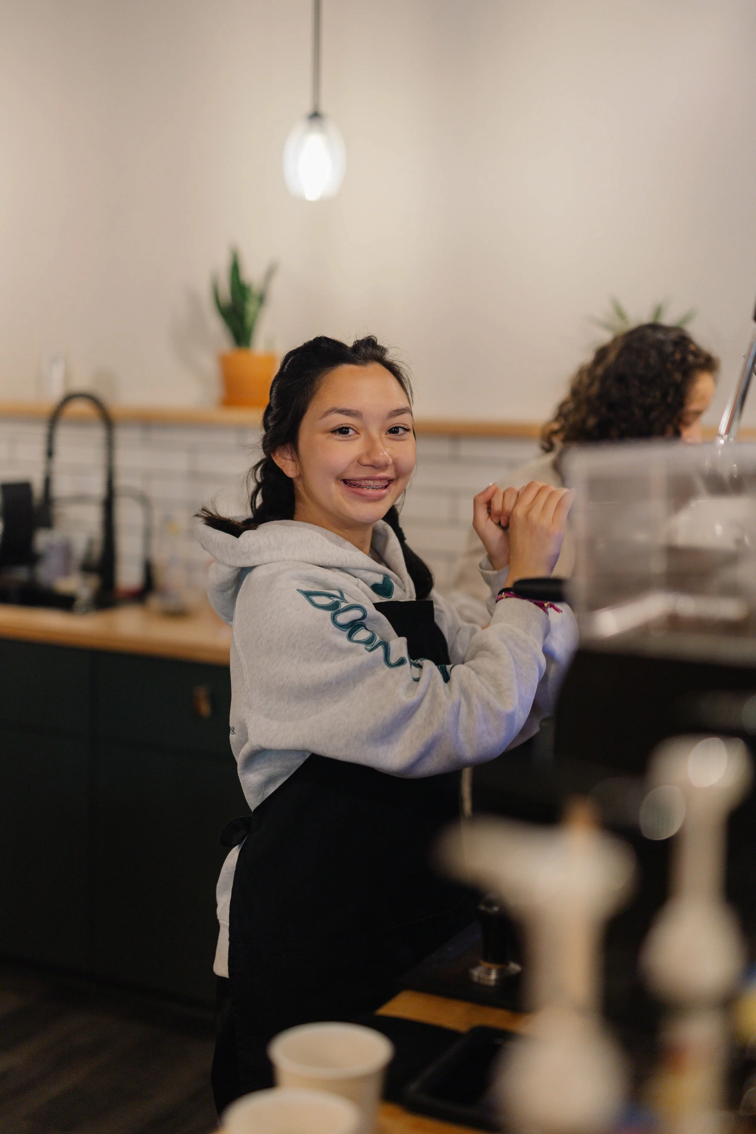 A young woman with dark hair styled in two braids smiling and wearing a gray hoodie with writing on one sleeve, standing in a cafe or kitchen with a blurred background, some plants, and various kitchen items.