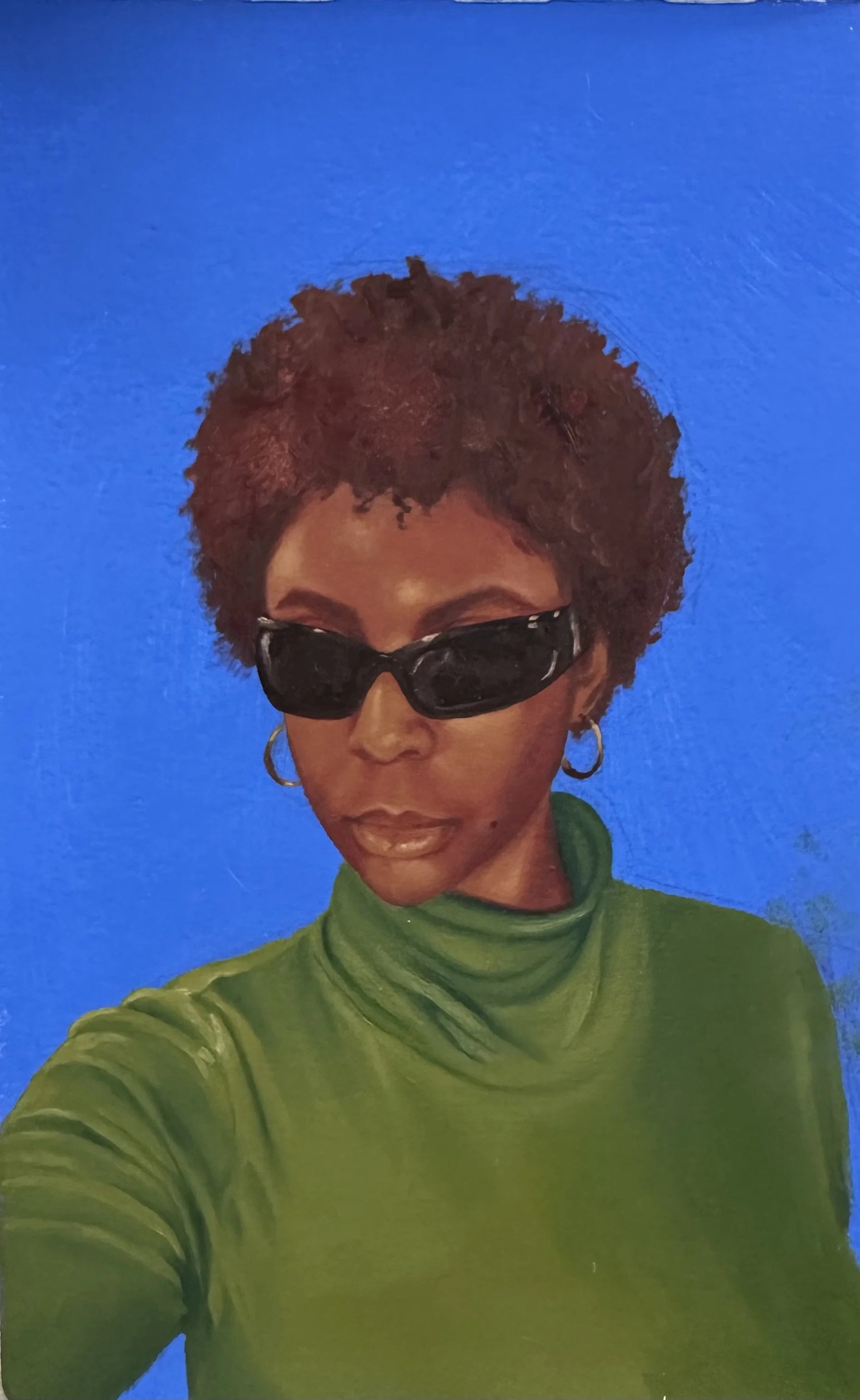 A woman with short curly hair wearing large black sunglasses, hoop earrings, and a green turtleneck sweater, against a bright blue background.