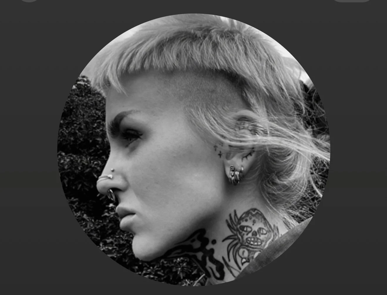 Black and white close-up profile of a woman with tattoos and multiple earrings, outdoors with trees in the background.