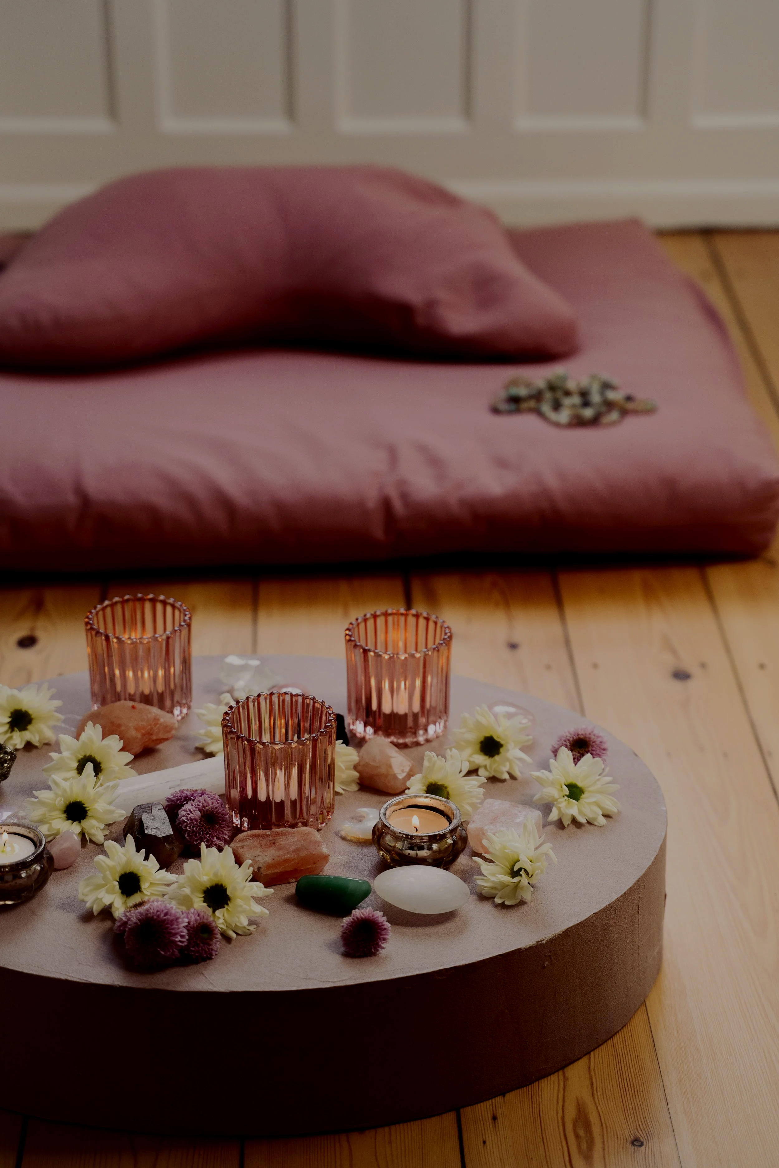 Decorative arrangement on a round platform with candles, flowers, and stones, in front of a pink cushion on a wooden floor.