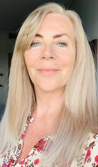 A close-up photo of a middle-aged woman with long blonde hair, blue eyes, and light skin, smiling slightly while looking at the camera. She is wearing a floral top and a small necklace, and the background is indoors with neutral colors.