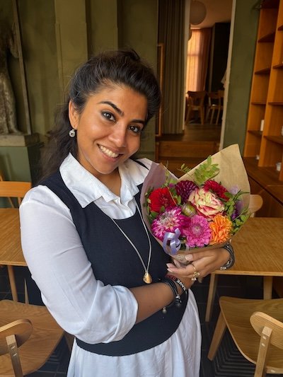 A woman with dark hair smiling and holding a bouquet of colorful flowers in a room with wooden furniture.