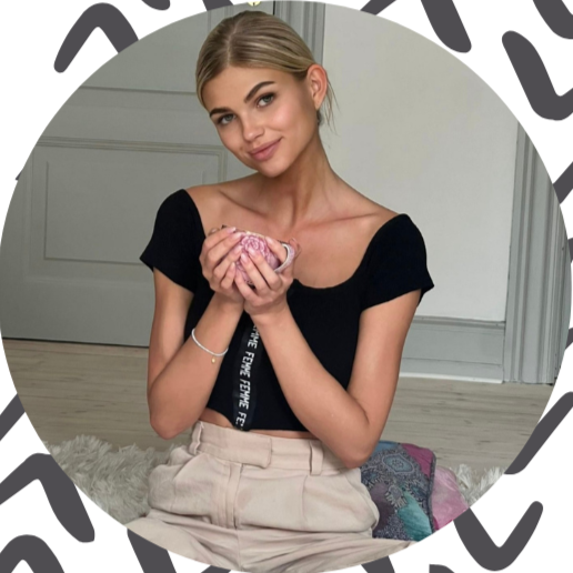 Girl sitting on a floor, holding a teacup with both hands, smiling at the camera, in a room with a gray door and patterned blanket.