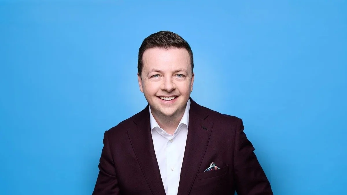 shows a photo of Oliver Callan, just his head and shoulders, smiling ay the camera, wearing a burgundy jacket with pocket square and white shirt on a sky blue background.