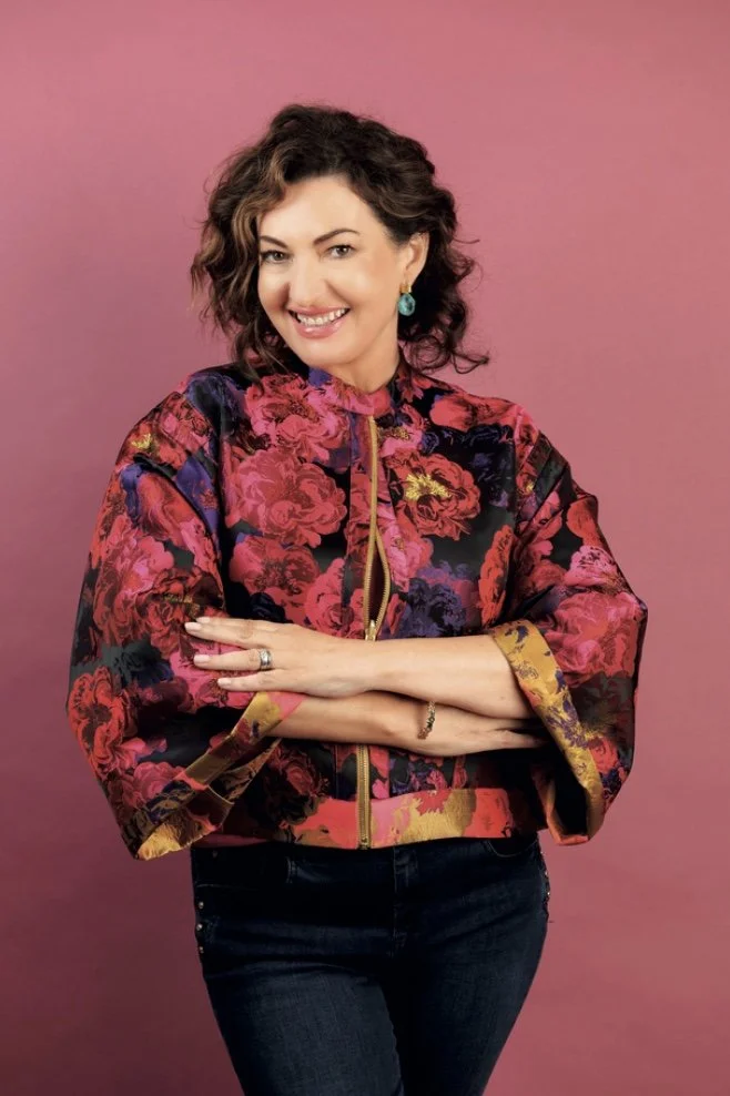 Maura Derrane smiling at the camera, wearing a floral wide sleeve zip top in pinks and purples, blue jeans and against a dusty pink background