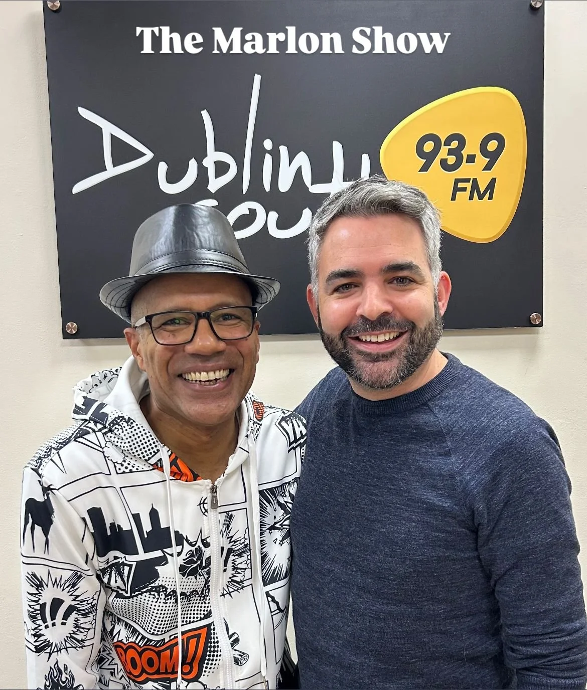 Shows Aidan & Marlon standing with an arm around each other smiling at the camera, with Dublin South 93.9 FM logo behind. Marlon wears a black leather hat and a white back and orange cartoon designed hoodie, while Aidan wears a navy sweater