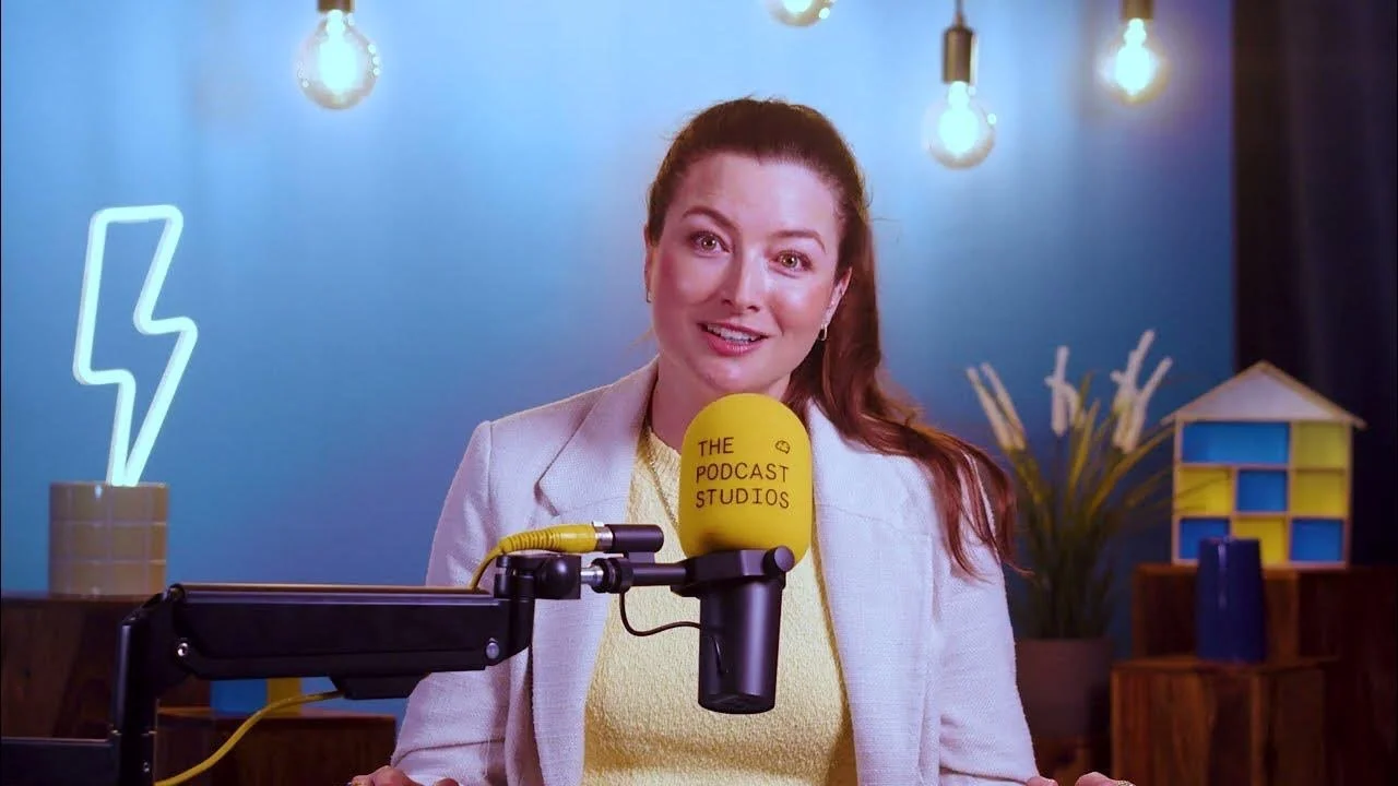 Meghann Scully sitting behind a spongey round yellow microphone that says the podcast studios. She wears a pale lemon top with a pastel pink jacket. her brown hair is tied back and following over her left shoulder.