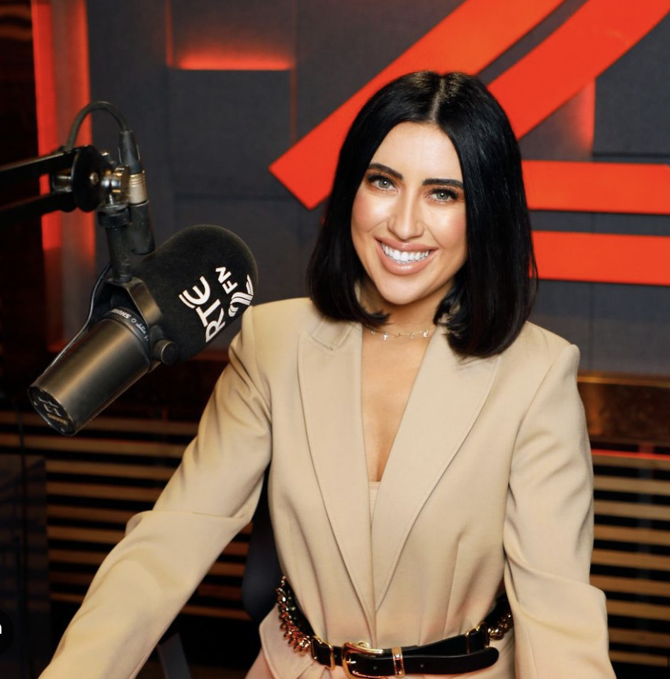 Lottie Ryan from 2fm smiling at the camera in a radio studio with a mic on her right, wearing a caramel coloured jacket with black and gold belt
