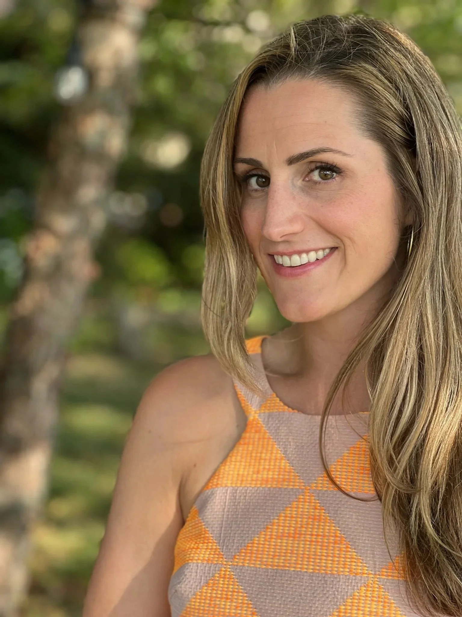 Patty Morrissey on the right hand side of he image, showing head and shoulders, blond shoulder length hair, tossed to her left, smiling directly at the camera wearing a sleeveless geometric patterned top in alternating triangles of grey and orange