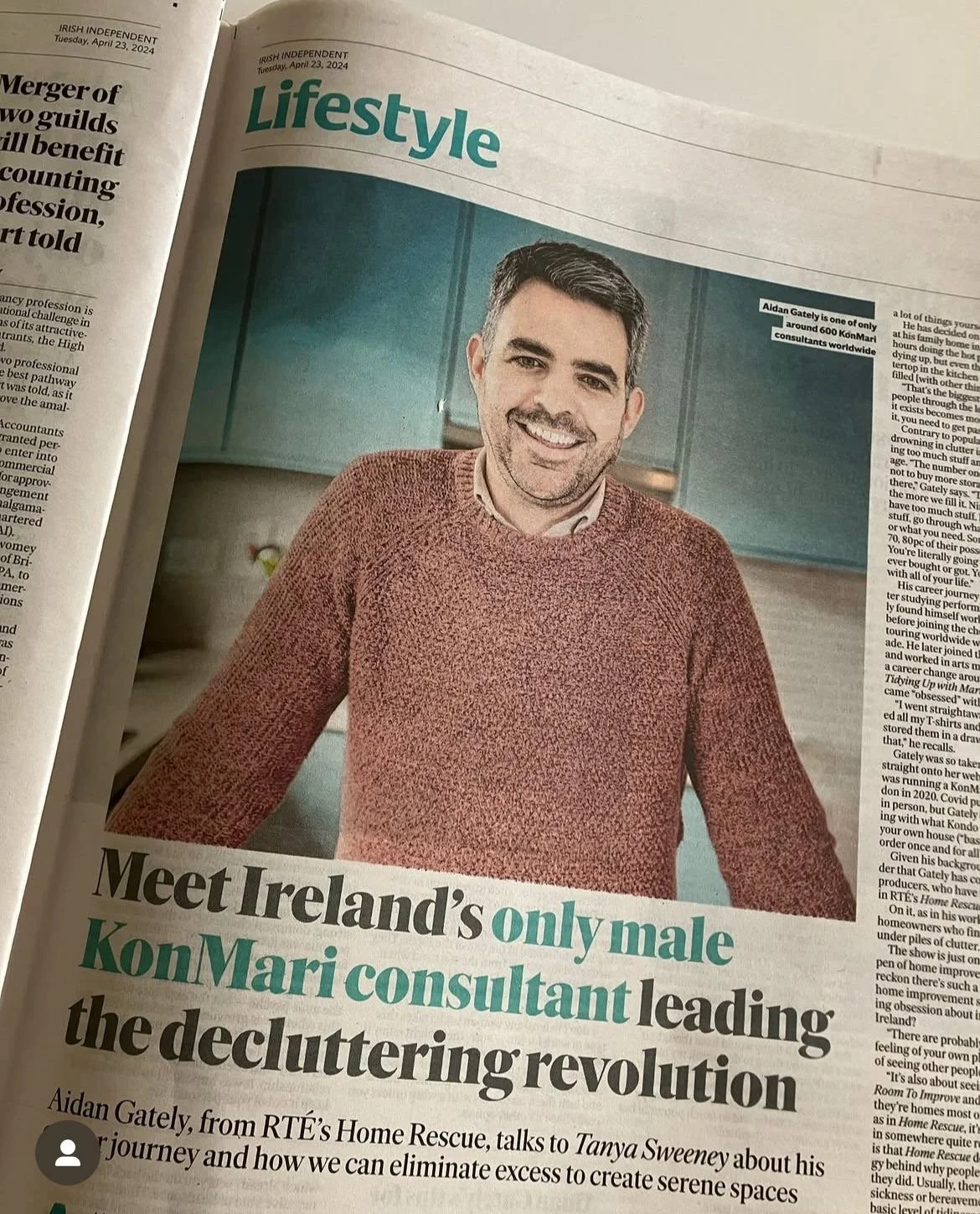 Shows Aidan Gately smiling at the camera, wearing a rust coloured sweater, he has some facial hair and black hair going grey. He's leaning on a counter in a light blue kitchen 