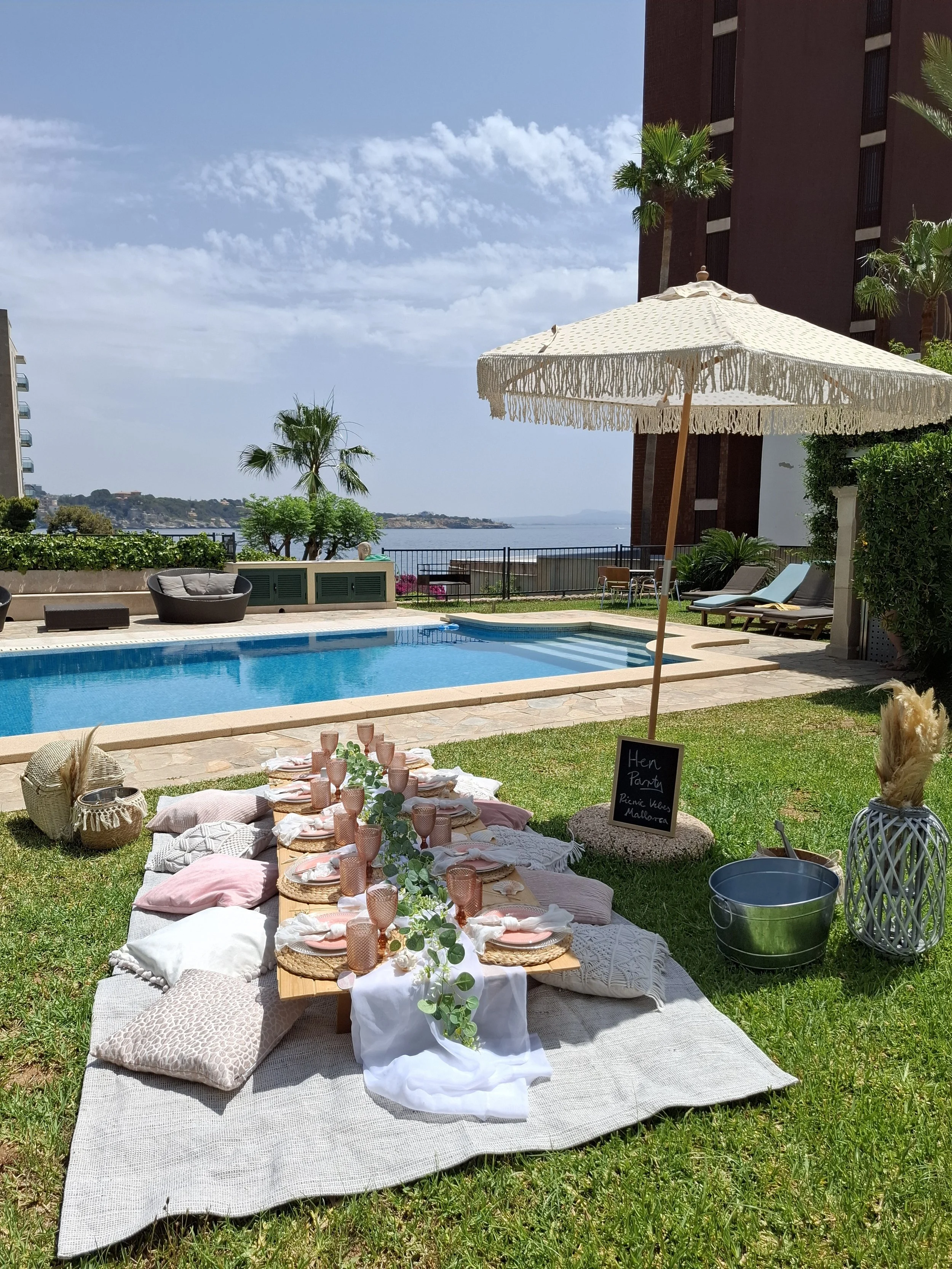 Beachside outdoor party setup with a picnic table, cushions, and a large umbrella. There is a pool in the background and a scenic view of the water and sky.