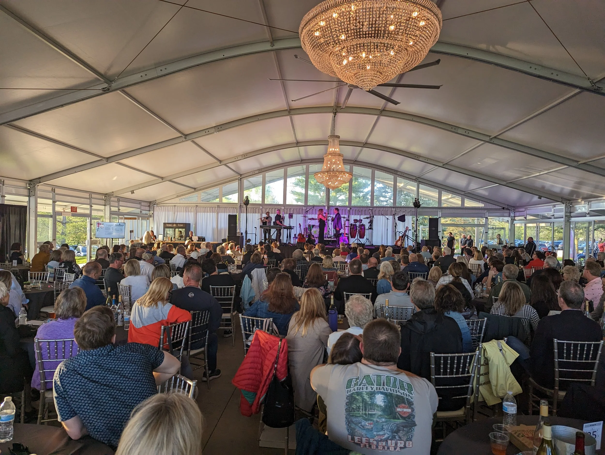 Indoor event with audience seated, people on stage, chandeliers, white tented ceiling.