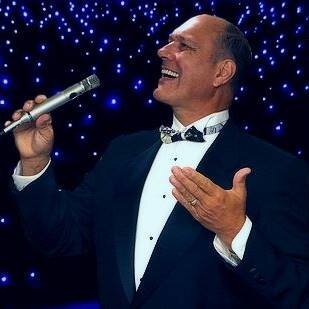 A man in a tuxedo singing into a microphone on a stage with blue starry background.