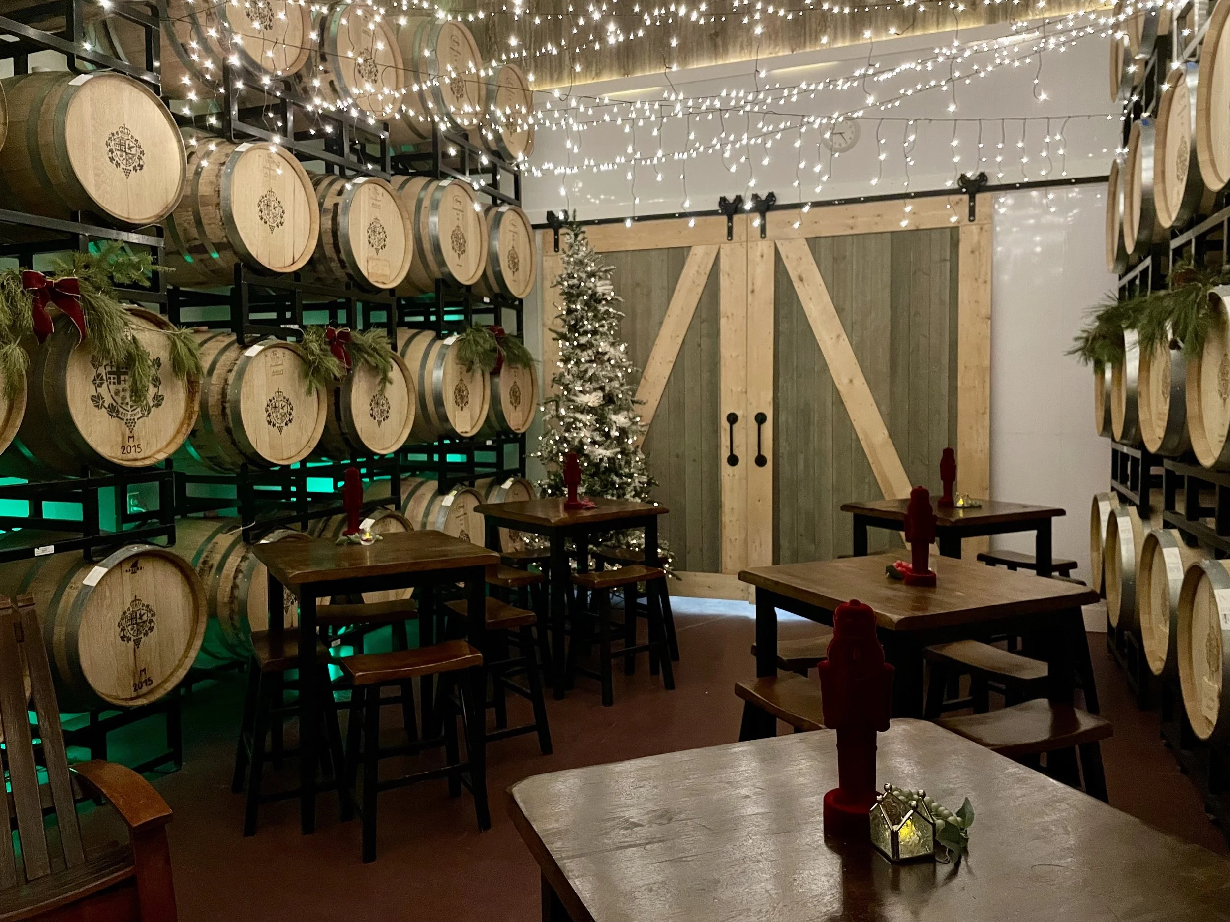 A room decorated for Christmas with string lights hanging from the ceiling, a small decorated Christmas tree, and wooden tables with festive candle holders and holiday decorations. There are shelves of wine barrels on the walls.