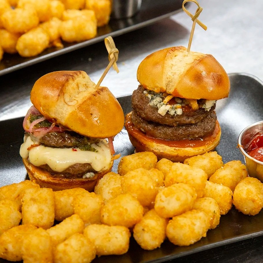 Two sliders with cheeseburger patties, toppings, and buns on a black tray with tater tots and ketchup.