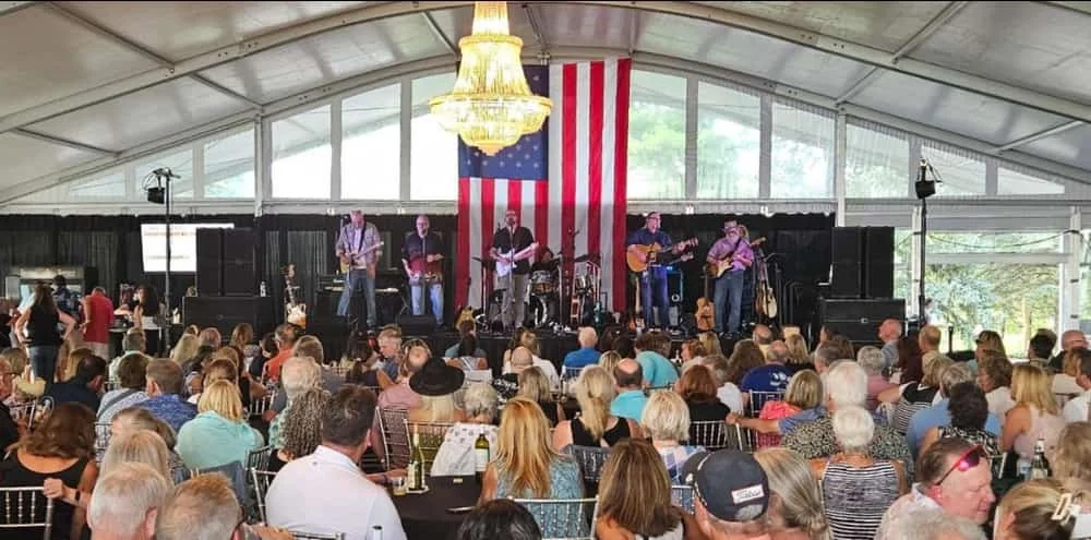A large indoor event with a live band performing on stage, decorated with an American flag backdrop and a chandelier, with many attendees seated at tables.