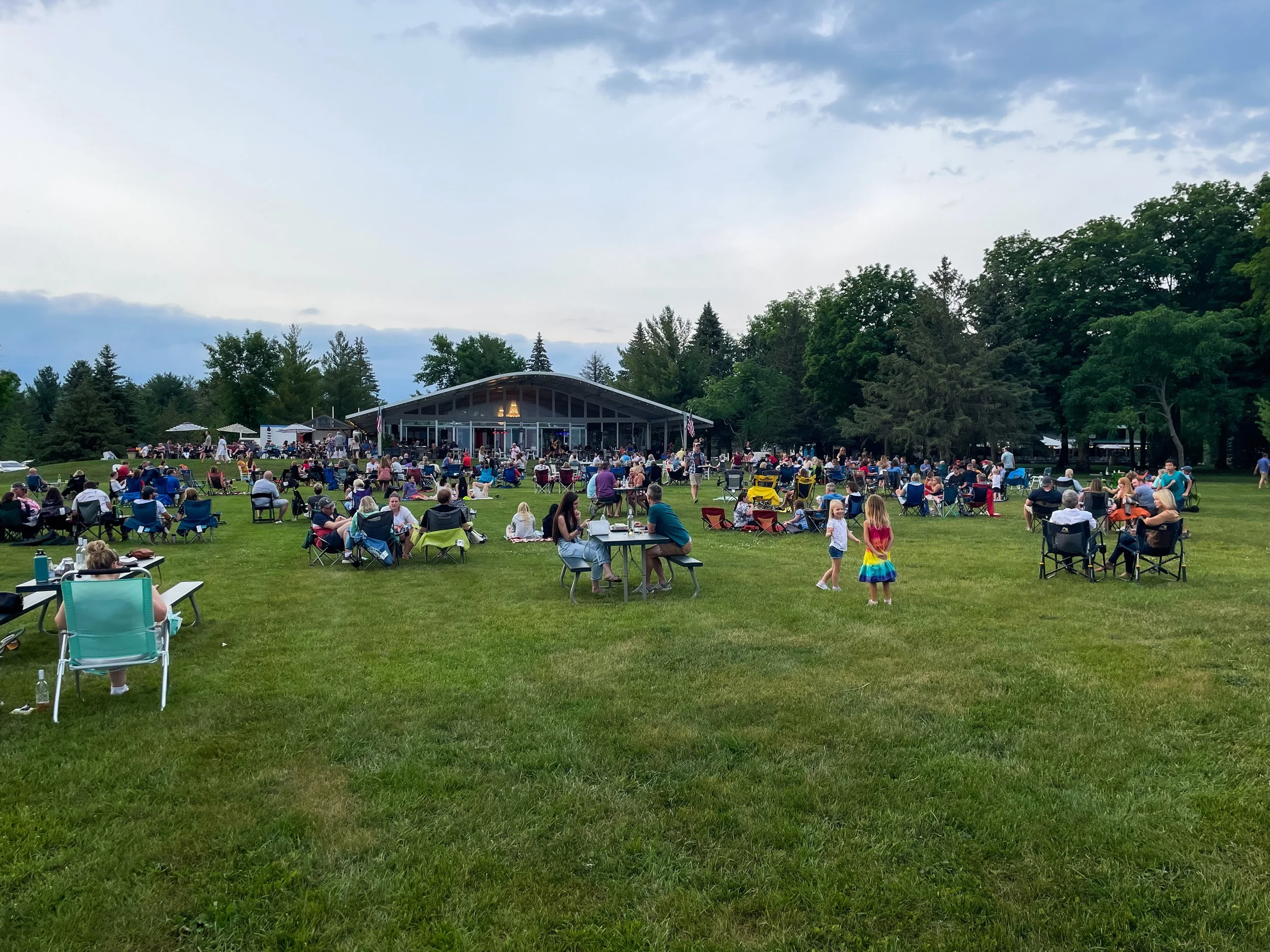 Outdoor gathering with people seated on a lawn in front of a pavilion, surrounded by trees and a cloudy sky.