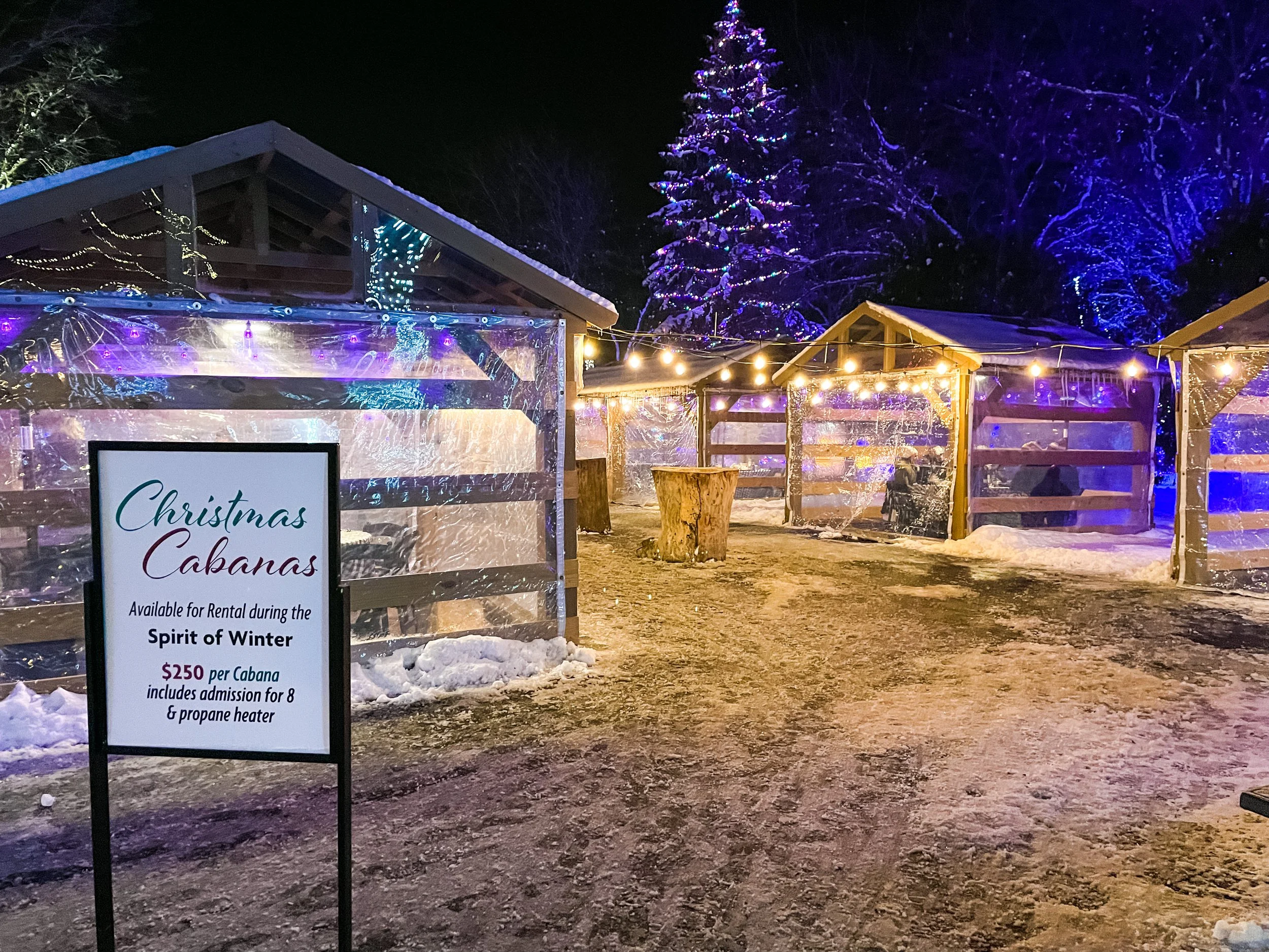 Festive outdoor Christmas cabanas decorated with string lights and surrounded by snow, with a sign promoting rental for winter gatherings.