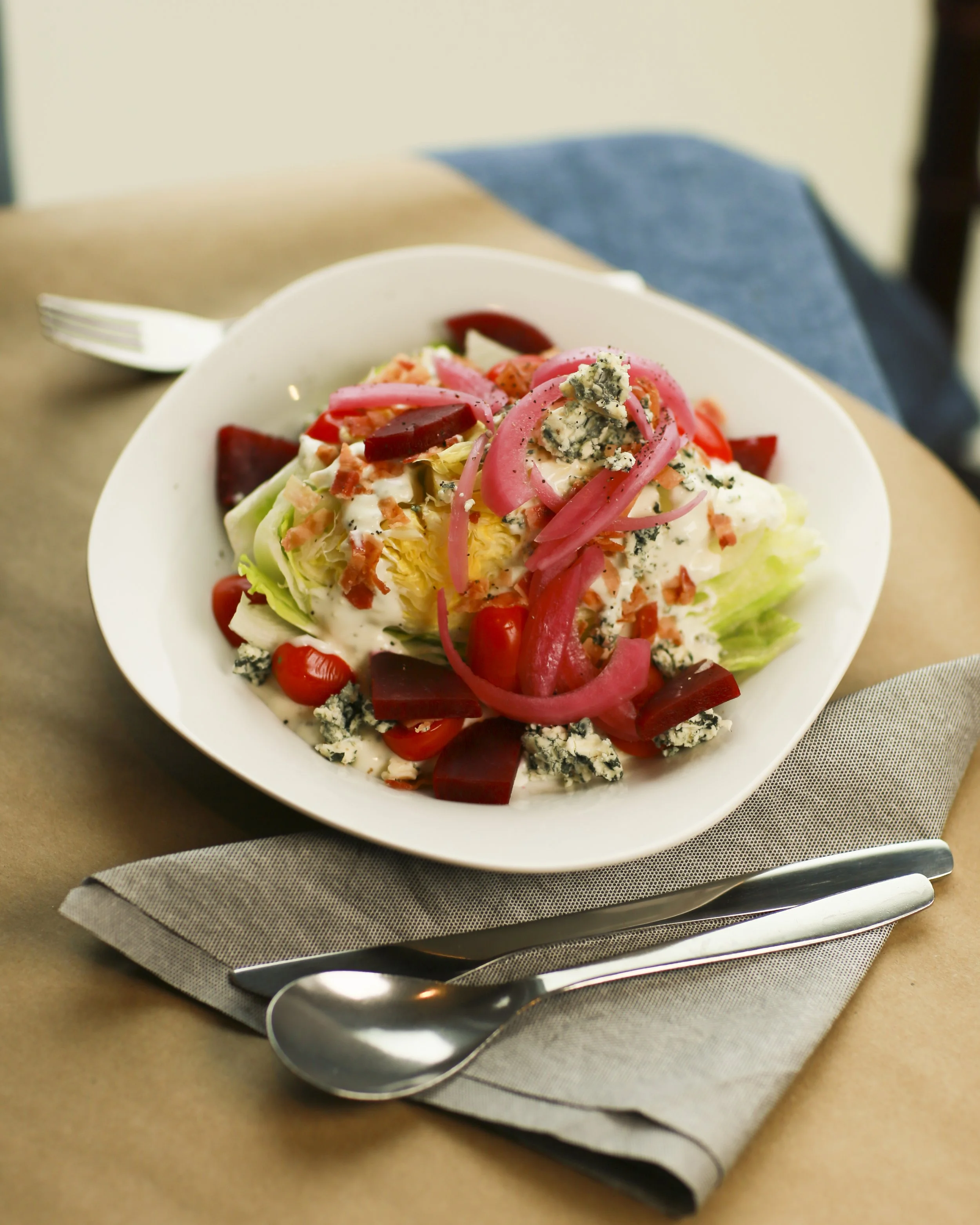 Lettuce salad with cherry tomatoes, blue cheese, pickled onions, bacon bits, and dressing in a white bowl on a napkin with a spoon and fork on a table.
