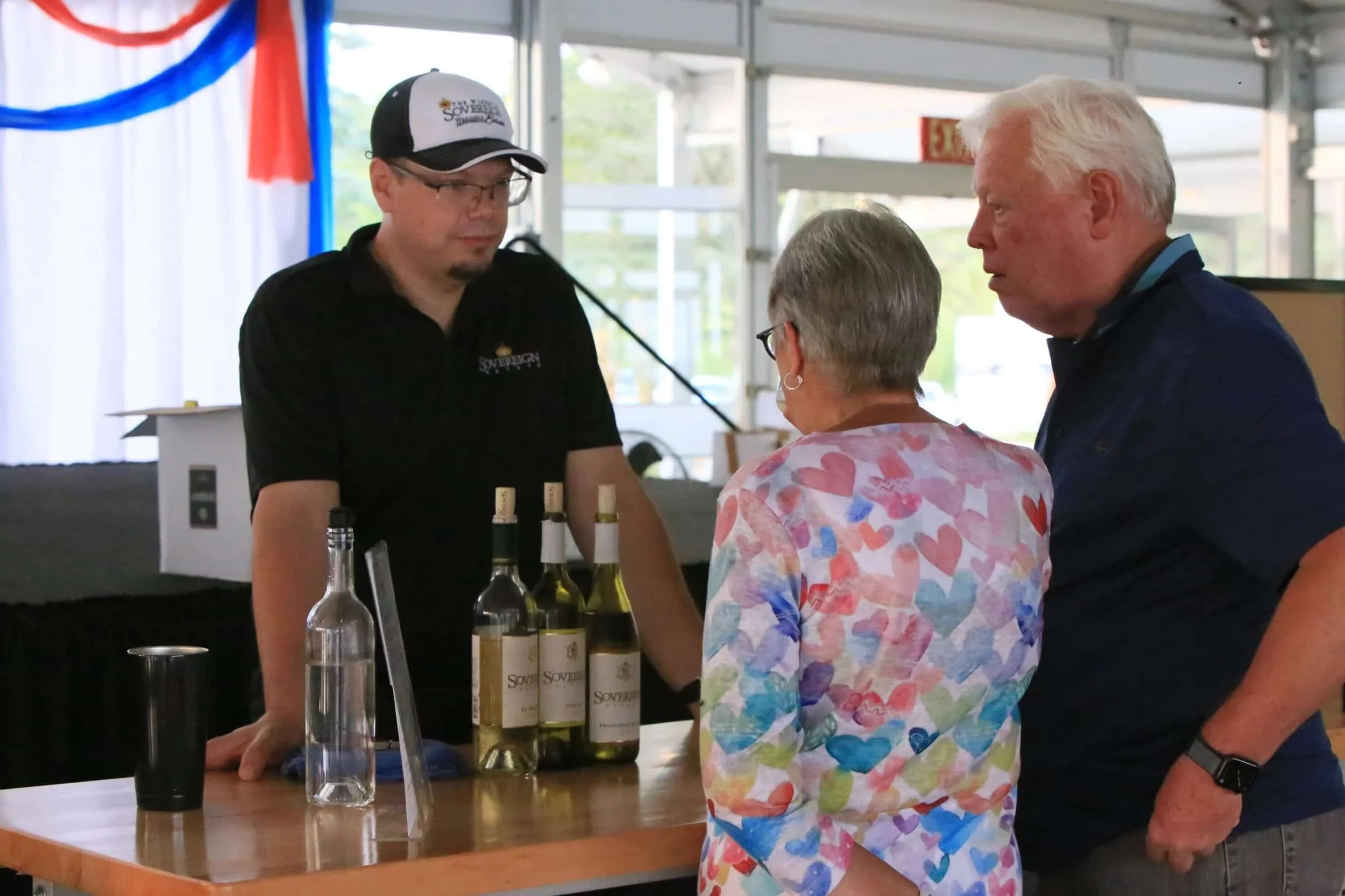 A man and a woman are talking to a person behind a counter with bottles of wine, inside a tent with a patriotic red, white, and blue decoration in the background.